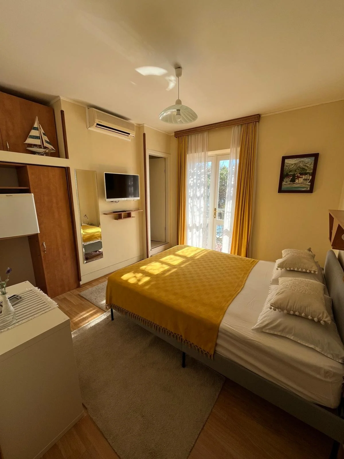 A cozy bedroom with a bed covered in white sheets and a yellow blanket, two pillows, a small rug, a wall-mounted TV, a mirror, and a window with yellow curtains letting in sunlight.