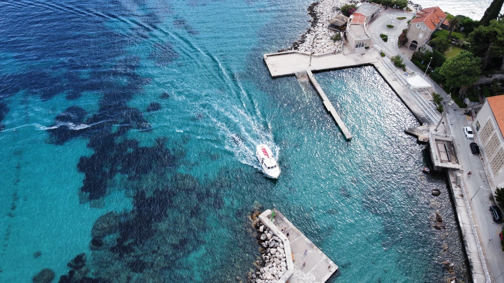 Aerial view of a marina with turquoise water, a small boat, a pier, and a coastal town with buildings, trees, and a street.