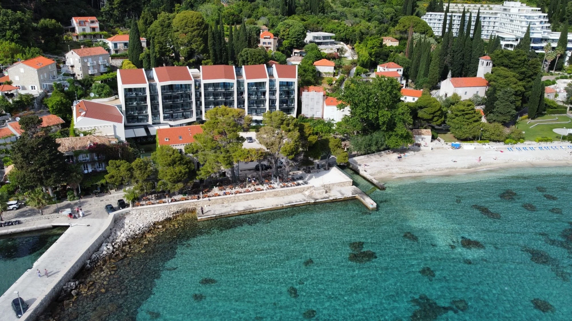Aerial view of a seaside area with a small beach, a pier, clear turquoise water, and a mix of modern and traditional buildings among green trees and hills.