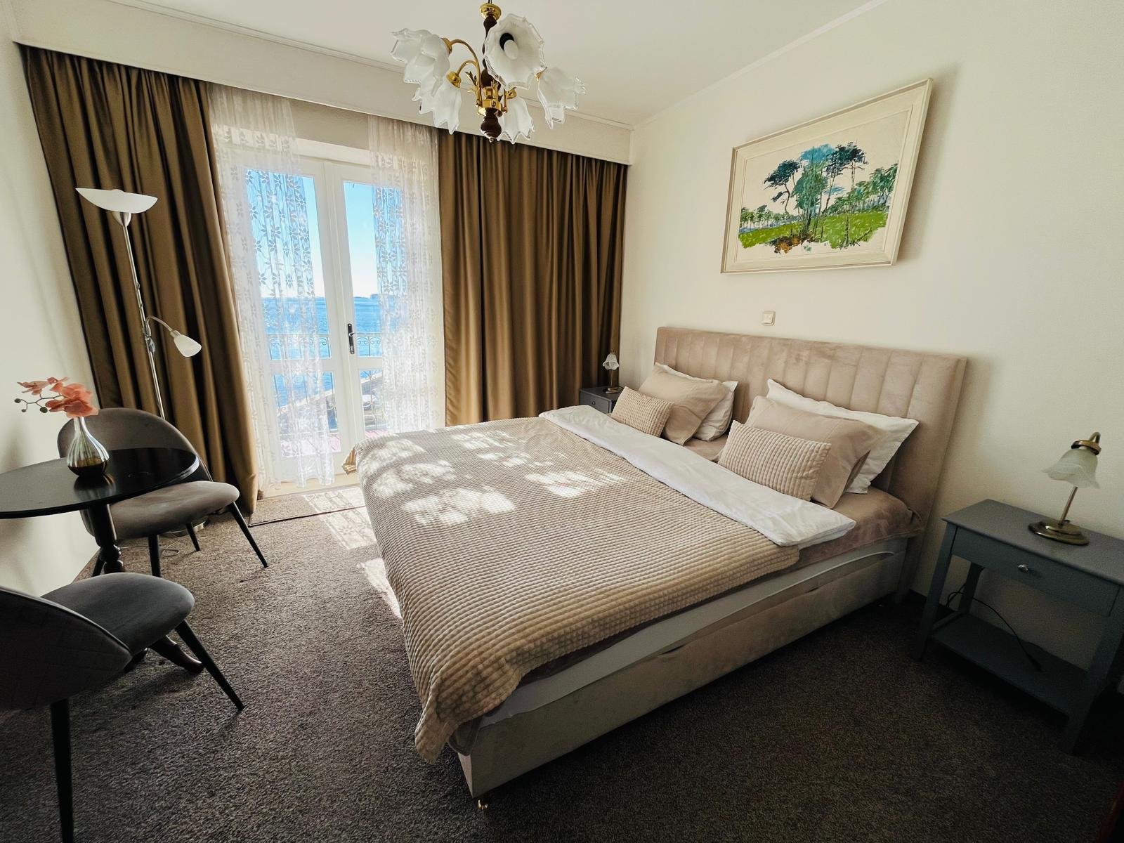 A hotel room with a large bed, beige headboard, and multiple pillows. Beside the bed are two gray nightstands with lamps. A painting of a landscape hangs above the bed. There is a small table with two chairs, a vase with flowers, a floor lamp, and cu