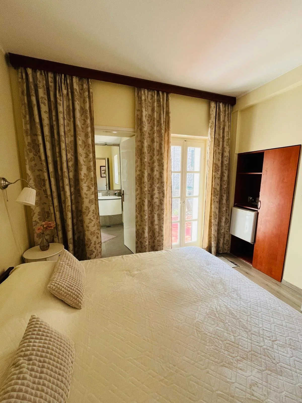 A cozy hotel room with a neatly made bed, a small side table with a lamp and flower vase, a window with patterned curtains, and a door leading to a bathroom. A small refrigerator is visible in a wooden cabinet.