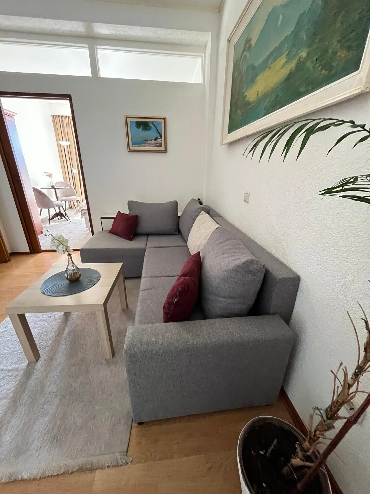 Living room with gray sectional sofa with red and beige cushions, a wooden coffee table with a vase, artwork on white walls, a doorway to a dining area with chairs and a table, and a potted plant.