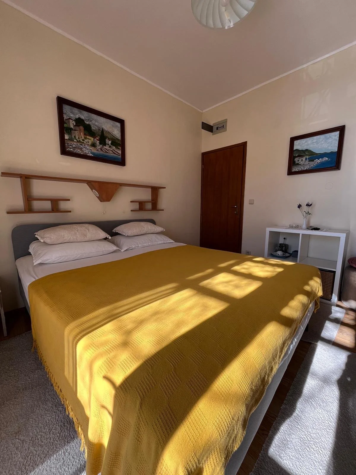 A bedroom with a large bed covered with a yellow blanket, white pillows, a wooden headboard with shelves, two framed landscape paintings on the walls, a small white shelf with decor, and sunlight casting shadows on the bed.