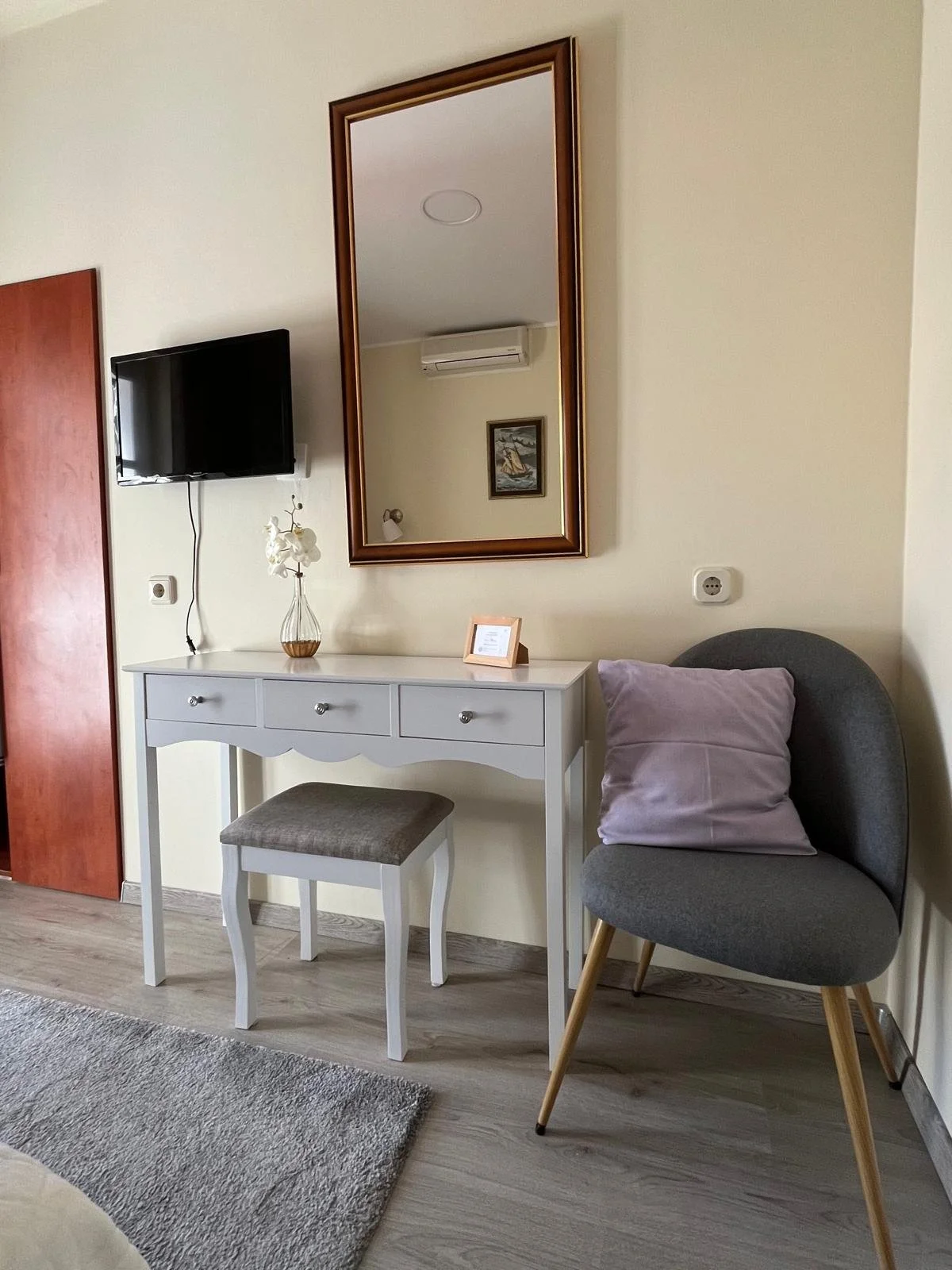 A hotel room corner with a white console table, a small upholstered stool, a modern gray armchair with a purple pillow, a large framed mirror, a small decorative vase with white flowers on the table, a wall-mounted TV, and a wall painting reflected i