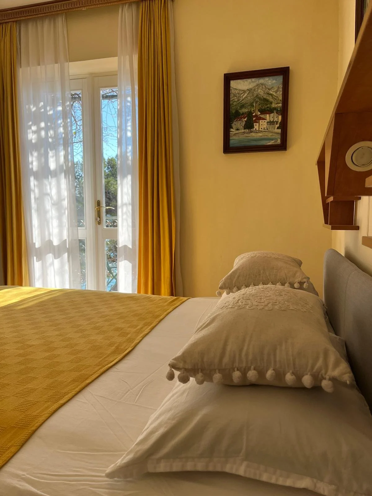 A neatly made bed with white sheets and beige pillows with pom-pom trimming, a yellow bed runner, and a view of a balcony with yellow and white curtains, a painting of mountains hangs on the wall.