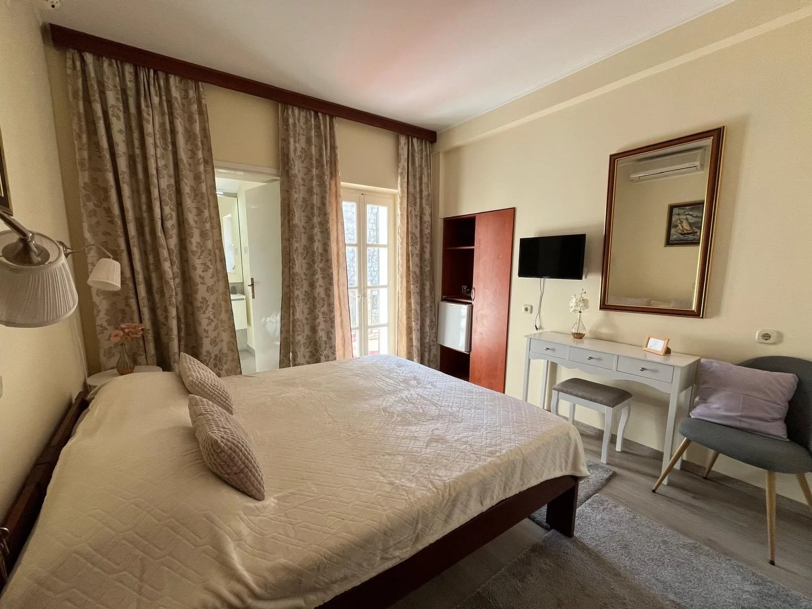 A cozy bedroom with a large bed covered in a beige quilt and two pillows, a bedside lamp, a white dresser with a mirror above it, a gray chair with a purple pillow, a wall-mounted flat-screen TV, and a door leading to a balcony with floral curtains, natural light, and a view of a balcony door.