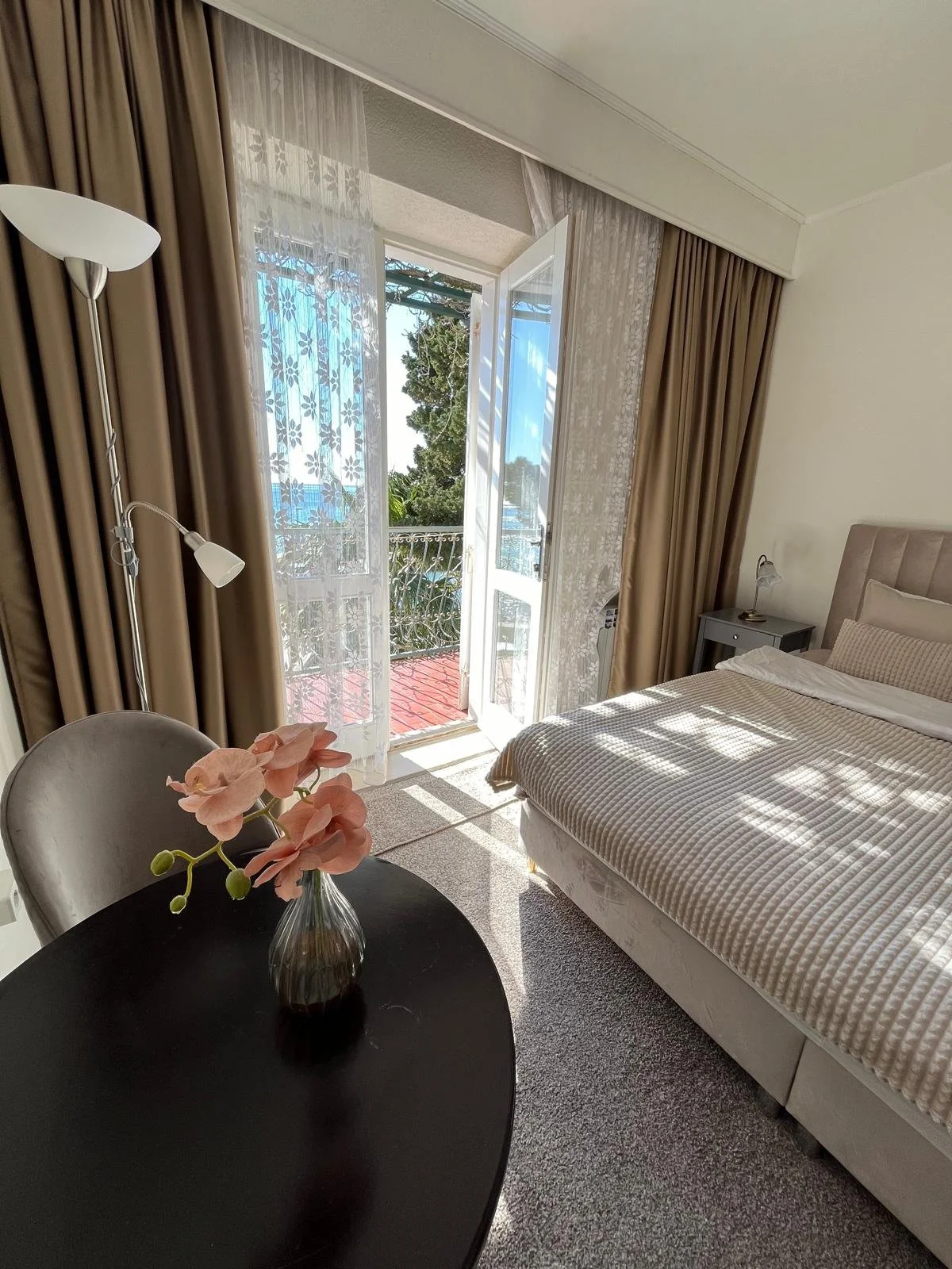 A cozy hotel room with a bed, a nightstand, and a black table with a pink flower vase. The room has large windows with sheer curtains and brown drapes, letting in sunlight and showing a balcony view of trees and a distant body of water.