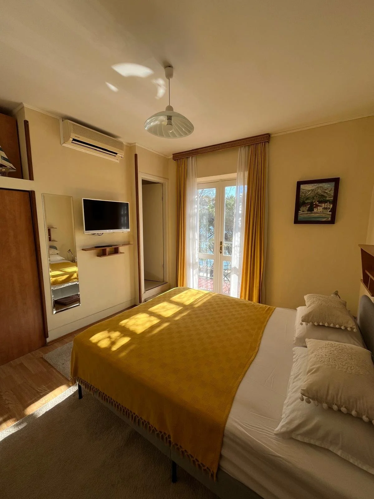 A cozy bedroom with a bed covered in a yellow blanket, pillows, a small window with yellow curtains, a wall-mounted TV, and sunlight streaming through the window.