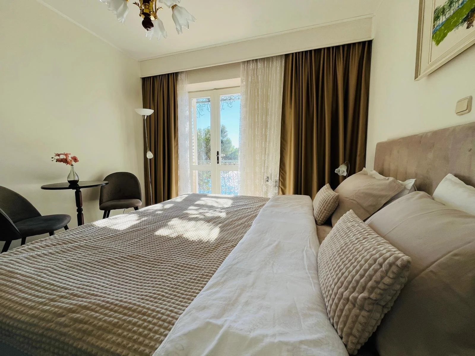 A cozy bedroom with a large bed, beige and white bedding, and multiple pillows, with sunlight streaming through a glass door with dark curtains, a small round table with two chairs, and a tall floor lamp in a bright room.