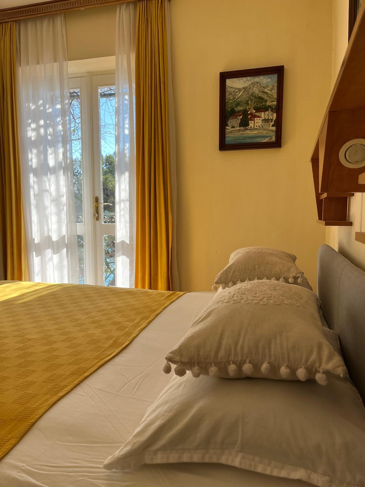 A bedroom with a bed covered in white, yellow, and beige linens, including decorative pillows with pom-pom borders, next to a window with white sheer curtains and yellow drapes. A framed painting of a mountain landscape hangs on the wall above the bed.