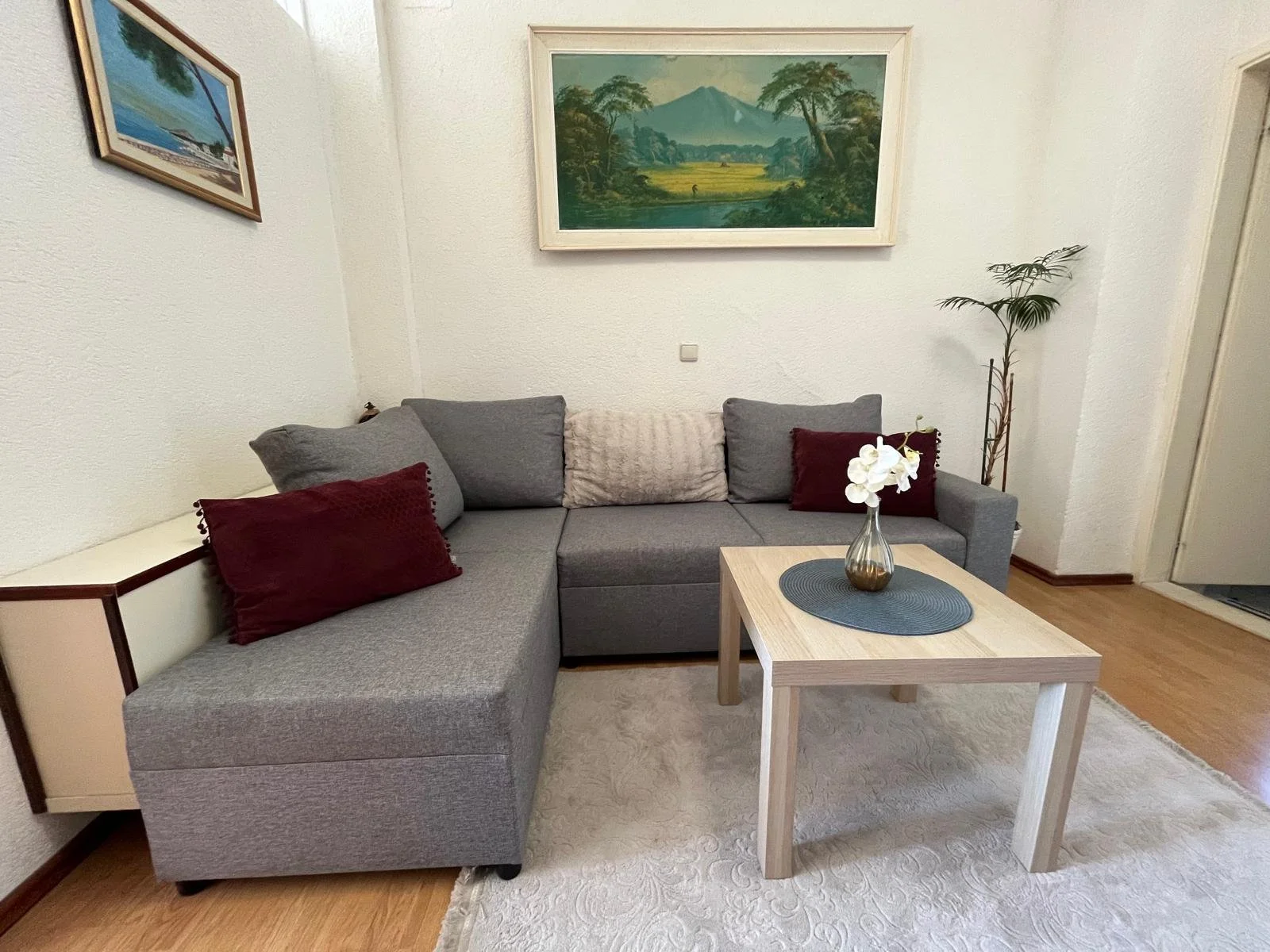 Living room with gray L-shaped sofa, red and gray throw pillows, a small light wood coffee table with a vase of white orchids, a patterned white rug, a potted plant in the corner, and framed landscape paintings on the white walls.
