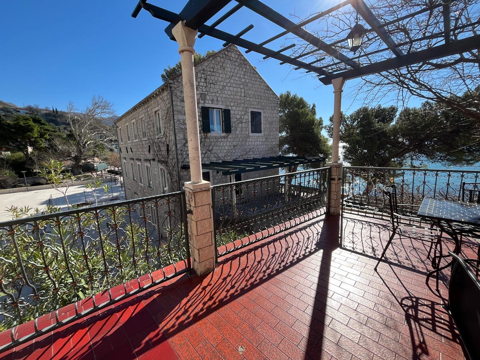 A balcony with black wrought iron railing and red tiled flooring, overlooking a stone building with black shutters, trees, and a clear blue sky.