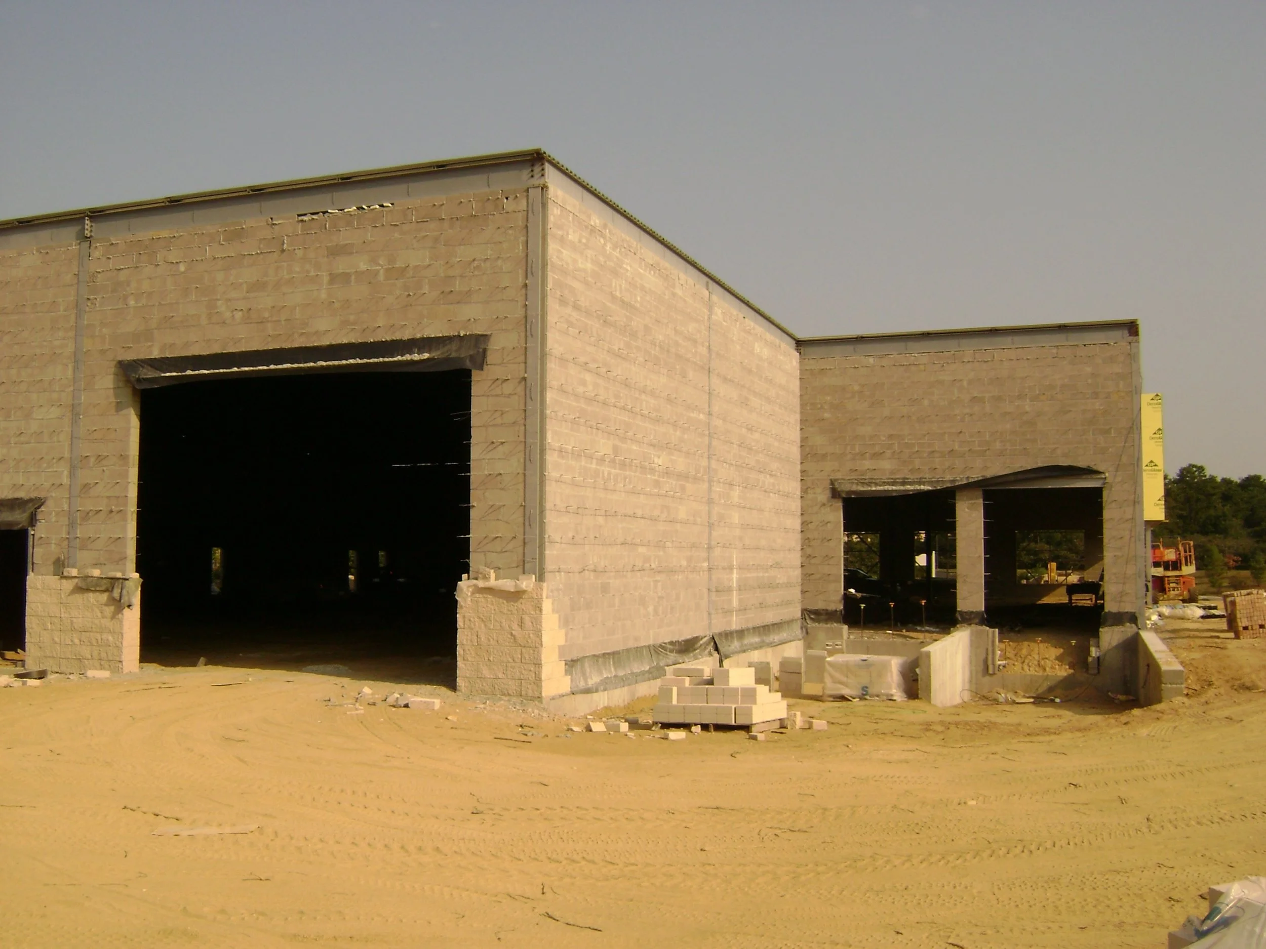 Under-construction building with large open garage doors, surrounded by sand and construction materials.