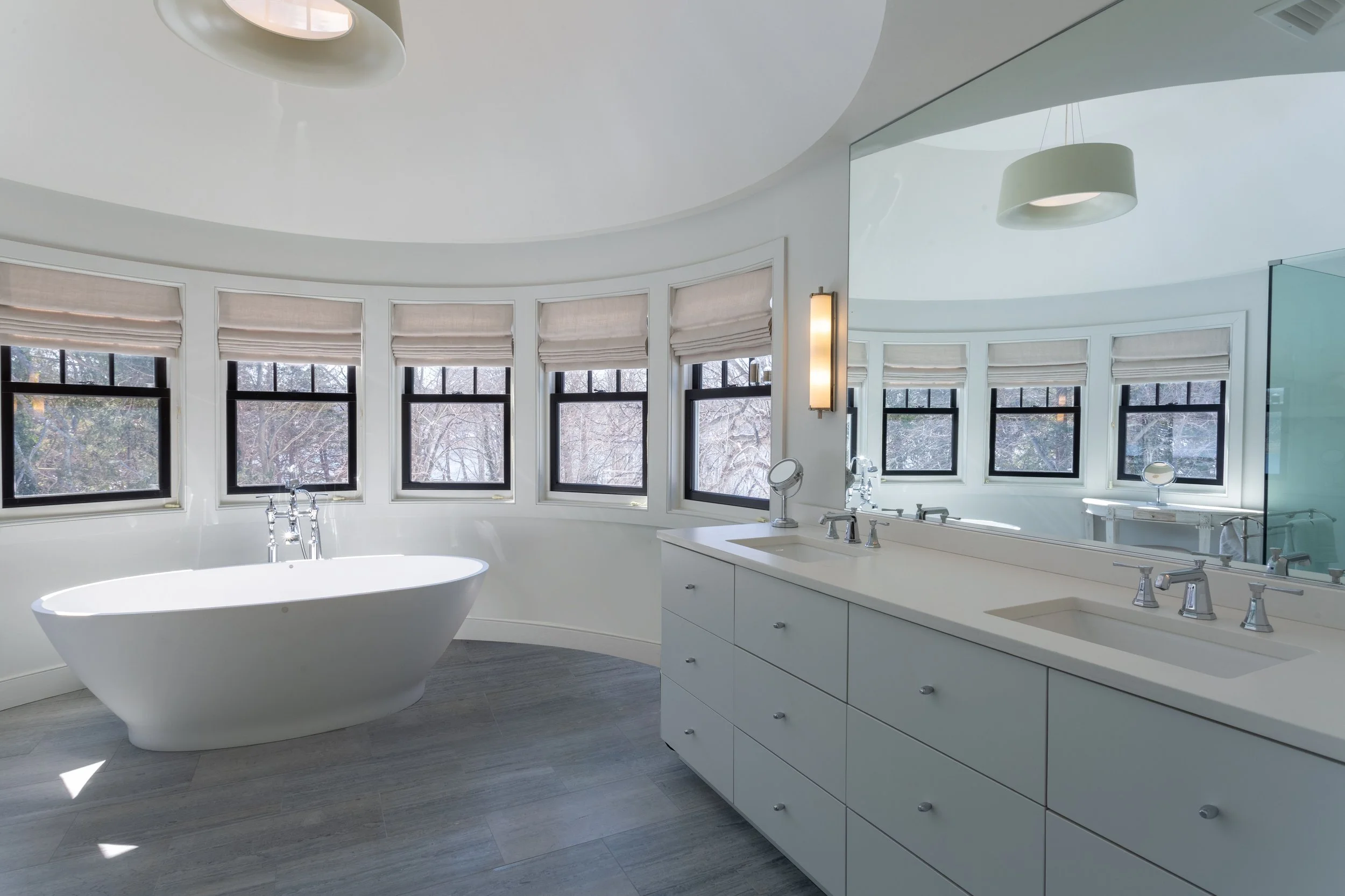 Modern bathroom with a freestanding bathtub below a row of six windows, and a wide mirror reflecting the room and a light fixture on the wall.