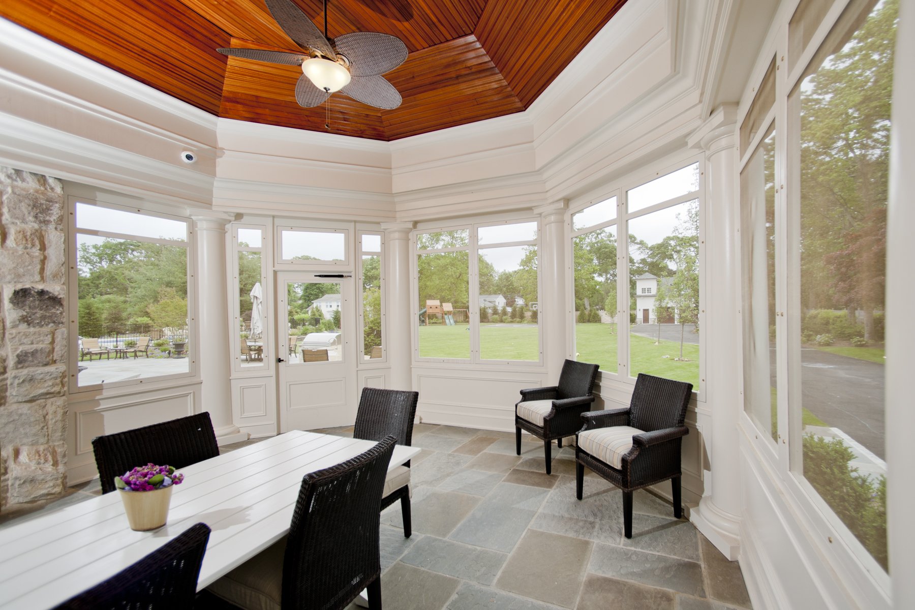 Sunroom with large windows, white walls, wicker chairs with cushions, a white table with a small potted plant, ceiling fan with leaf-shaped blades, stone wall section, and view of a green yard with trees and houses outside.