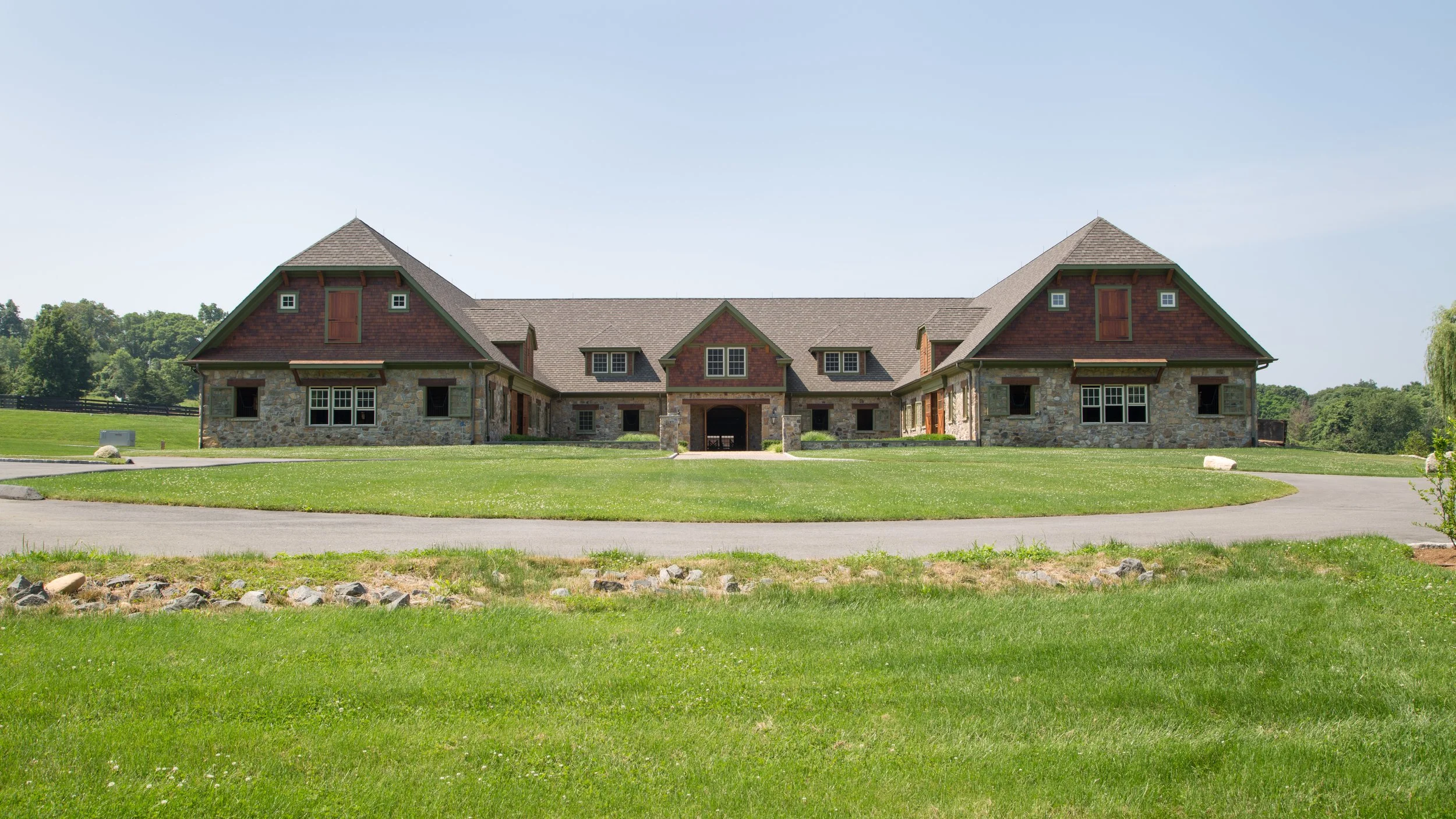 Large house with stone and wood exterior, multiple gabled roofs, set on a grassy lawn with a circular driveway, under a clear blue sky.