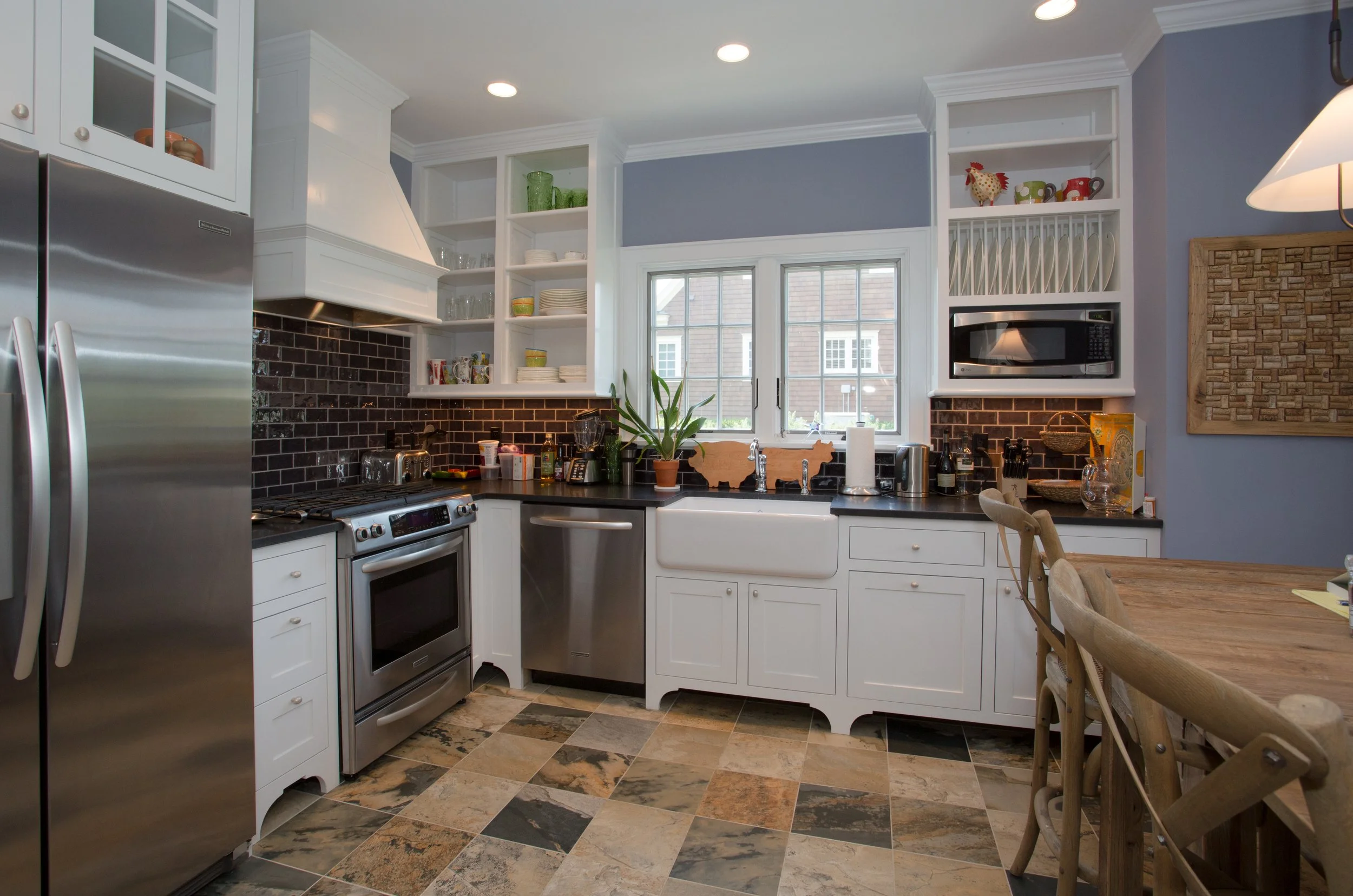 Kitchen with white cabinets, black countertops, black tile backsplash, stainless steel appliances, and a window above the sink. There are open shelves with dishes and mugs, a plant on the counter, and a wooden dining table with chairs.