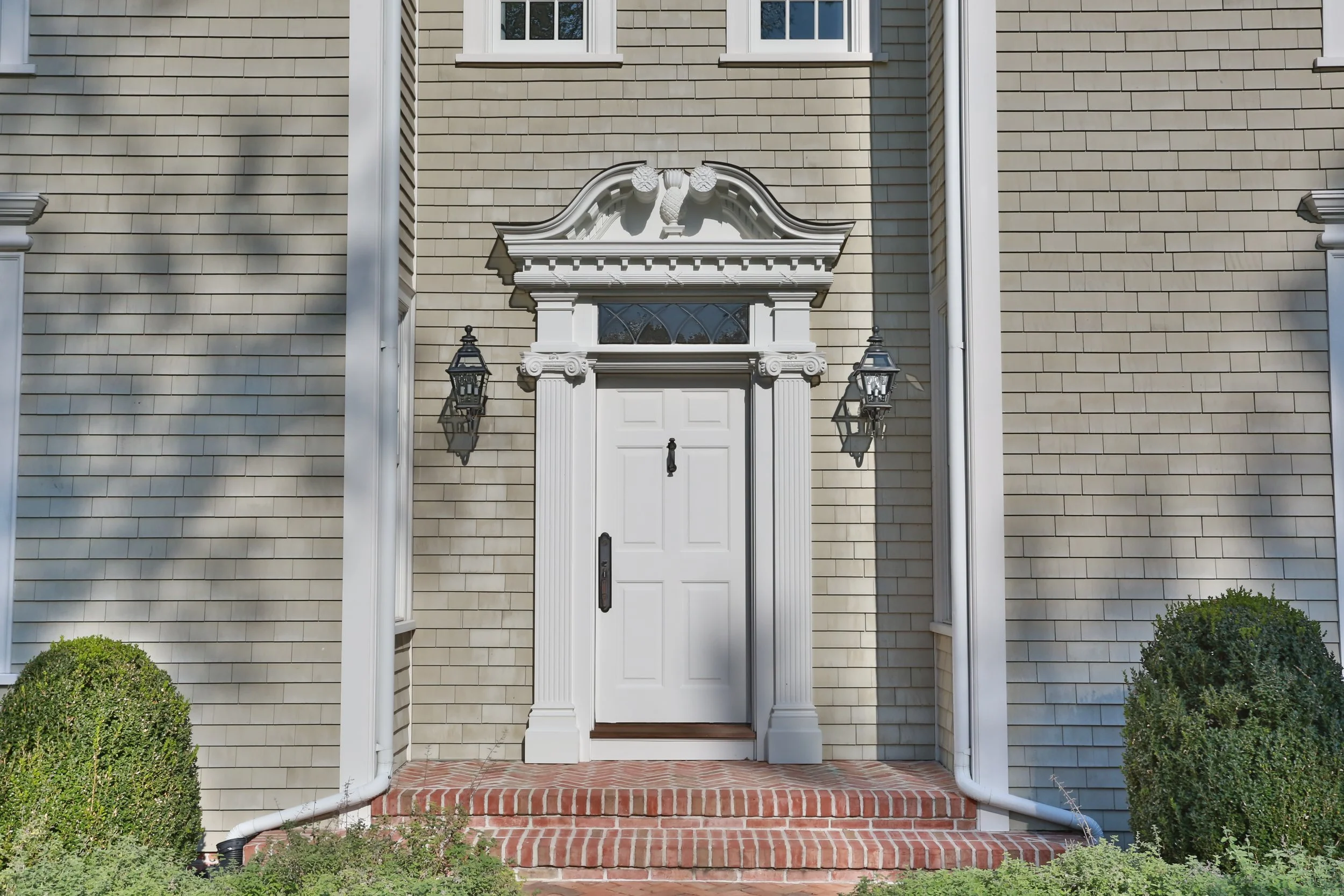 White front door with a decorative arched pediment and two black lantern-style wall sconces on a beige brick house with trimmed bushes in front.