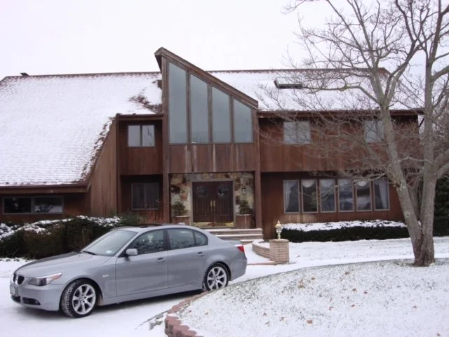 Wooden house with a sloped roof, large windows, and a prominent triangular glass section, surrounded by snow, with a silver car parked in front and a leafless tree to the right.