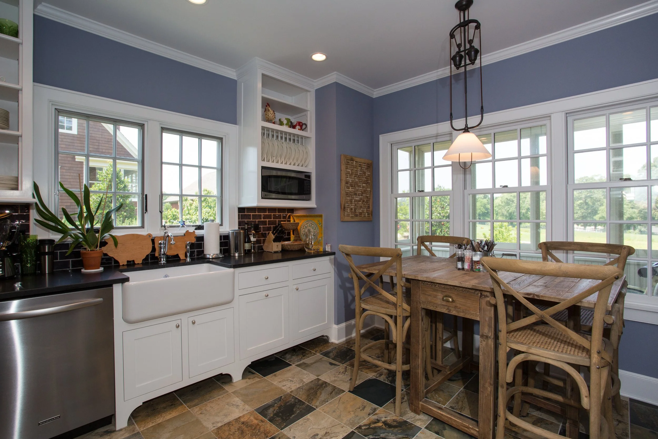 A bright kitchen with white cabinets, black countertops, a farmhouse sink, and large windows. There is a wooden dining table with four chairs and a hanging light fixture. Decor includes plants, dishes, and kitchen utensils.