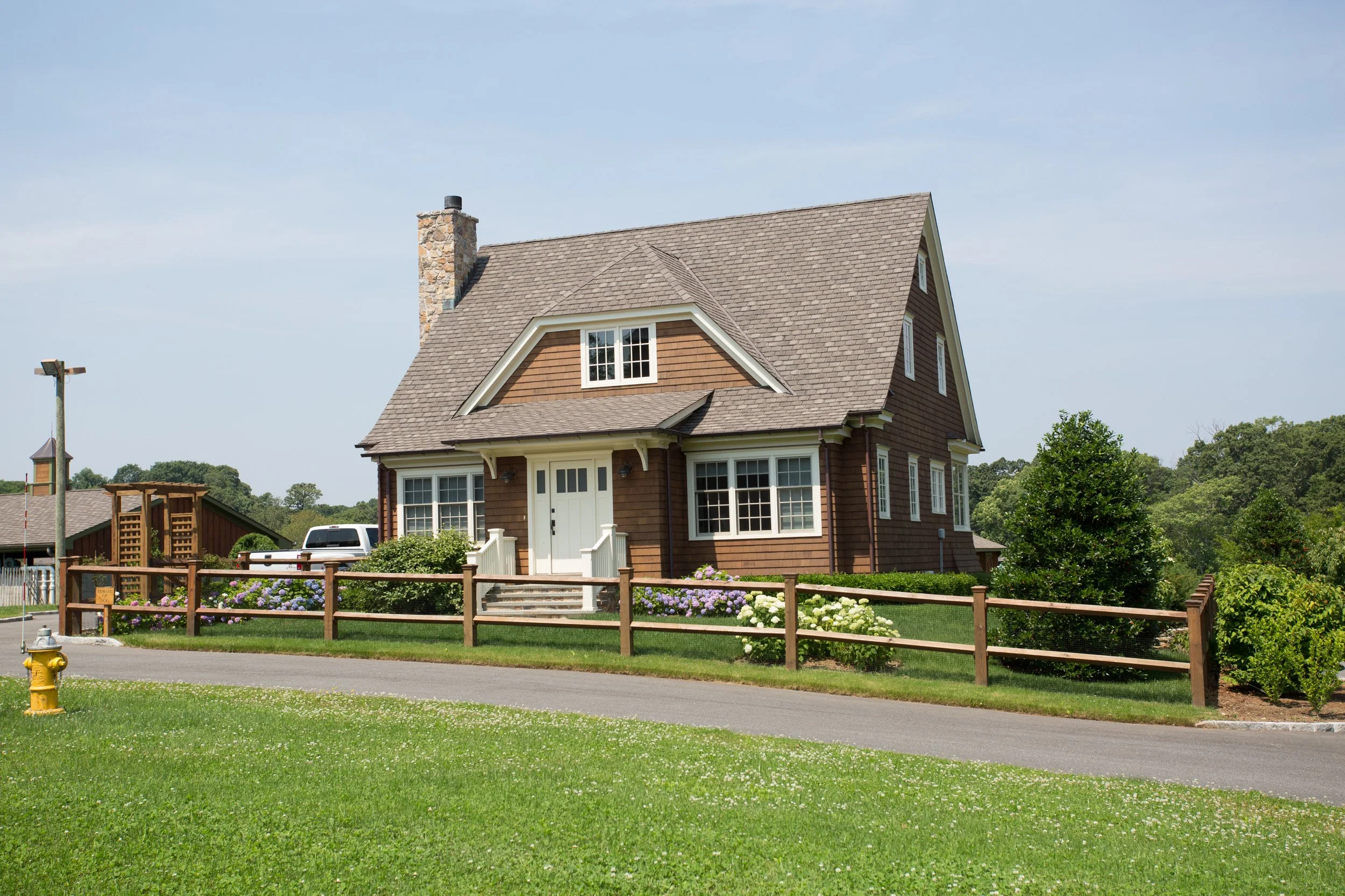 A two-story house with a gable roof, brown siding, and white trim, surrounded by a neatly maintained lawn and a small fence, with a driveway and trees in the background.