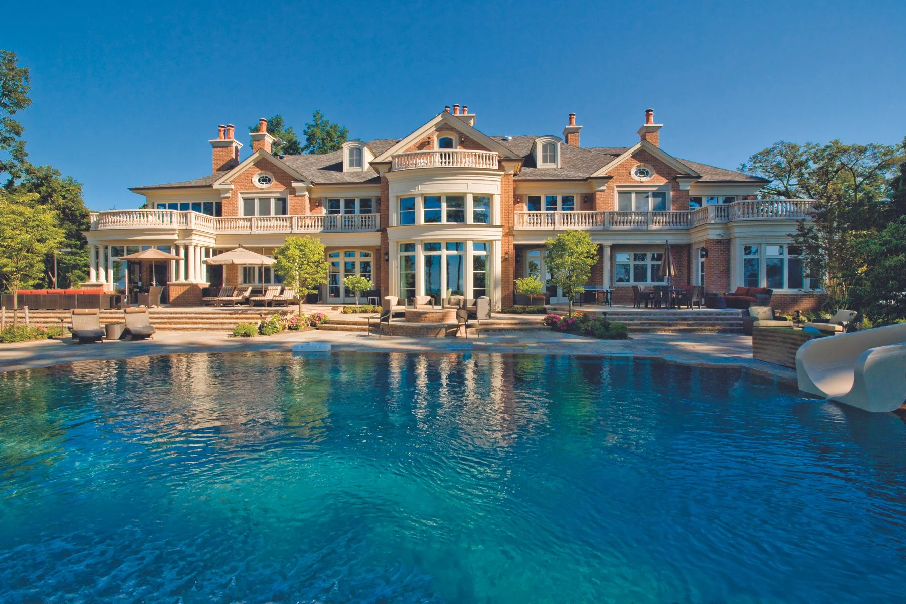 Large brick mansion with multiple stories, balconies, and large windows, set behind a swimming pool with lounge chairs and outdoor seating.