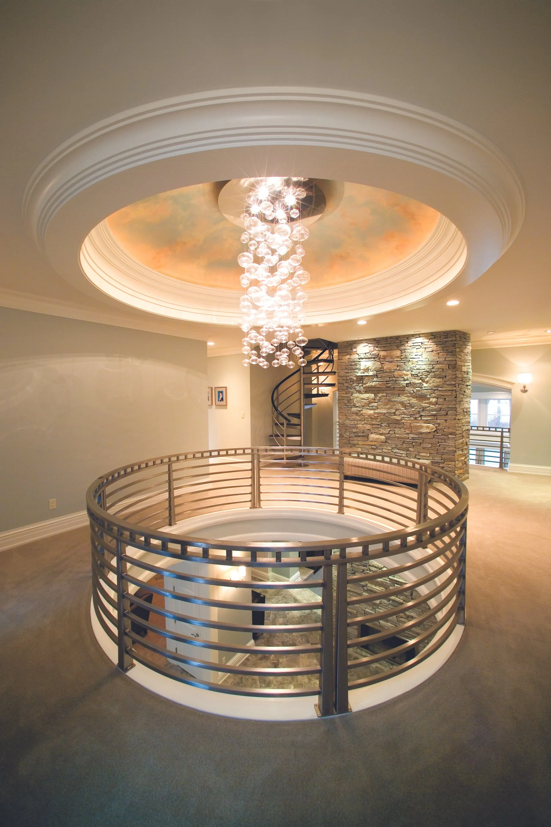 Interior of a modern multi-story house showcasing a chandelier, a painted ceiling, a spiral staircase, and a metal railing around an open staircase to the lower level, with stone wall accents and large windows.
