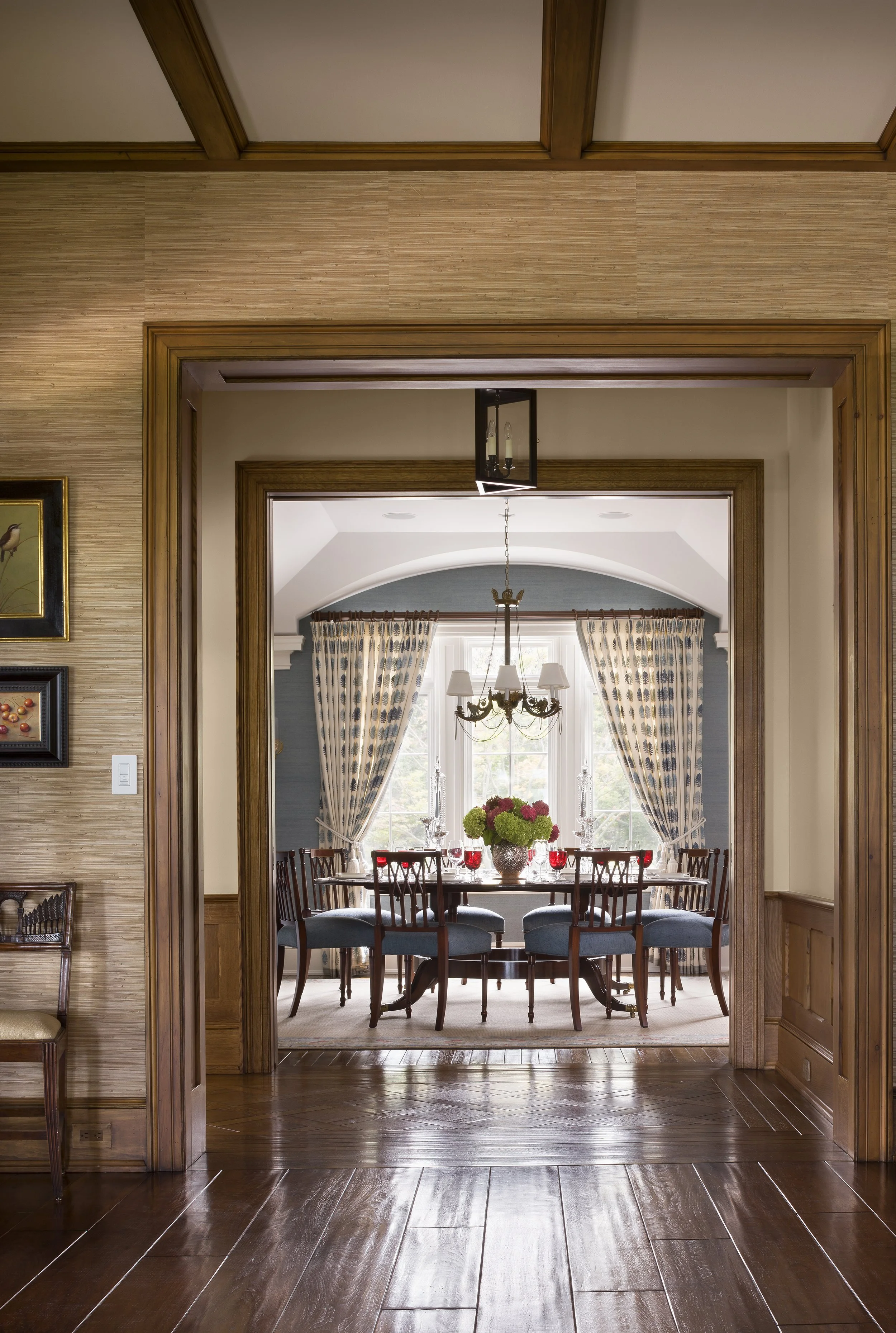 A view through a wooden doorway into a dining room with a window, blue patterned curtains, a chandelier, a round table set with red wine, glasses, a flower arrangement, and six chairs.