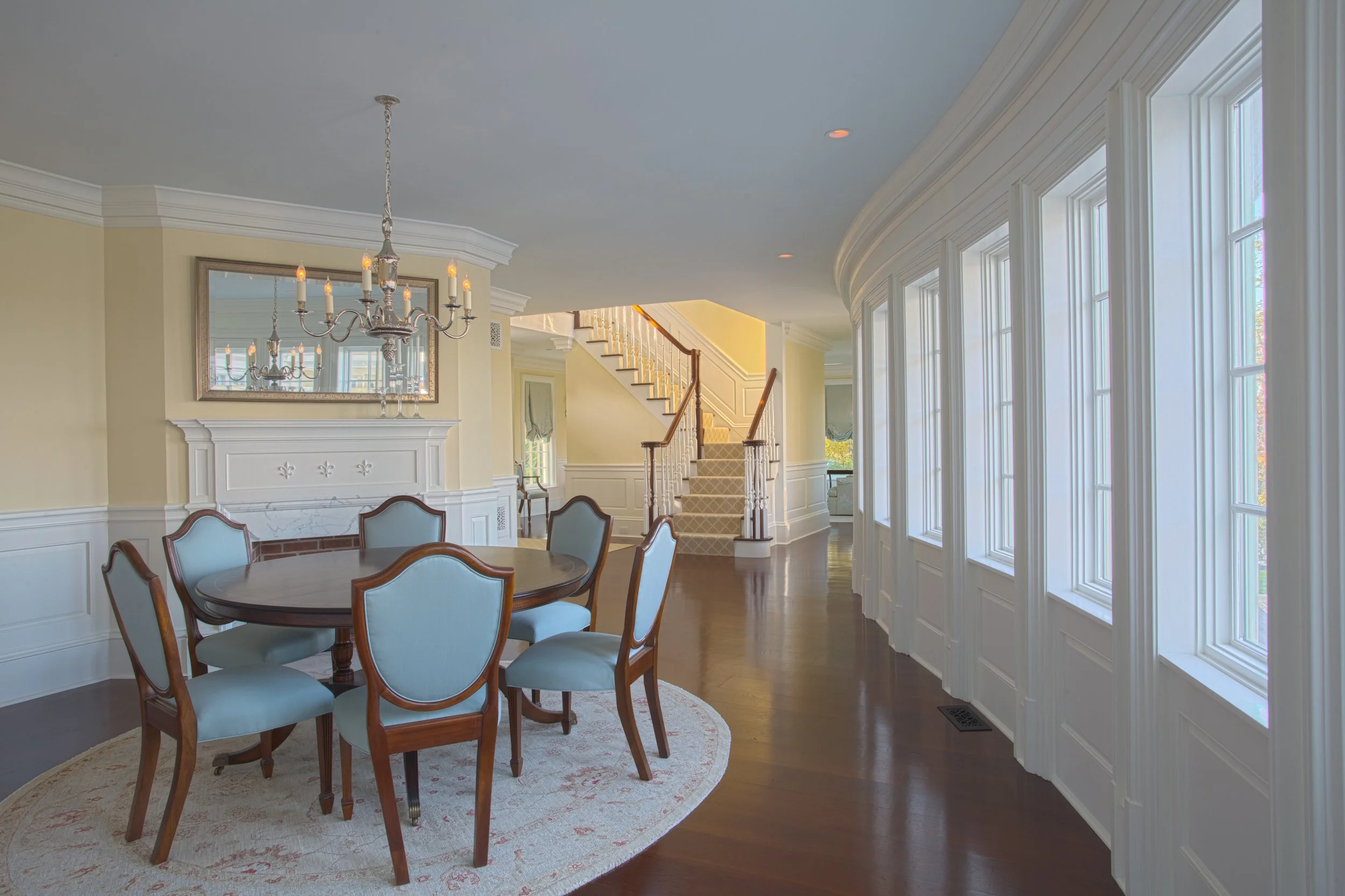 Elegant dining room with a round wooden table, light blue upholstered chairs, a chandelier, large windows with white trim, and a staircase in the background.