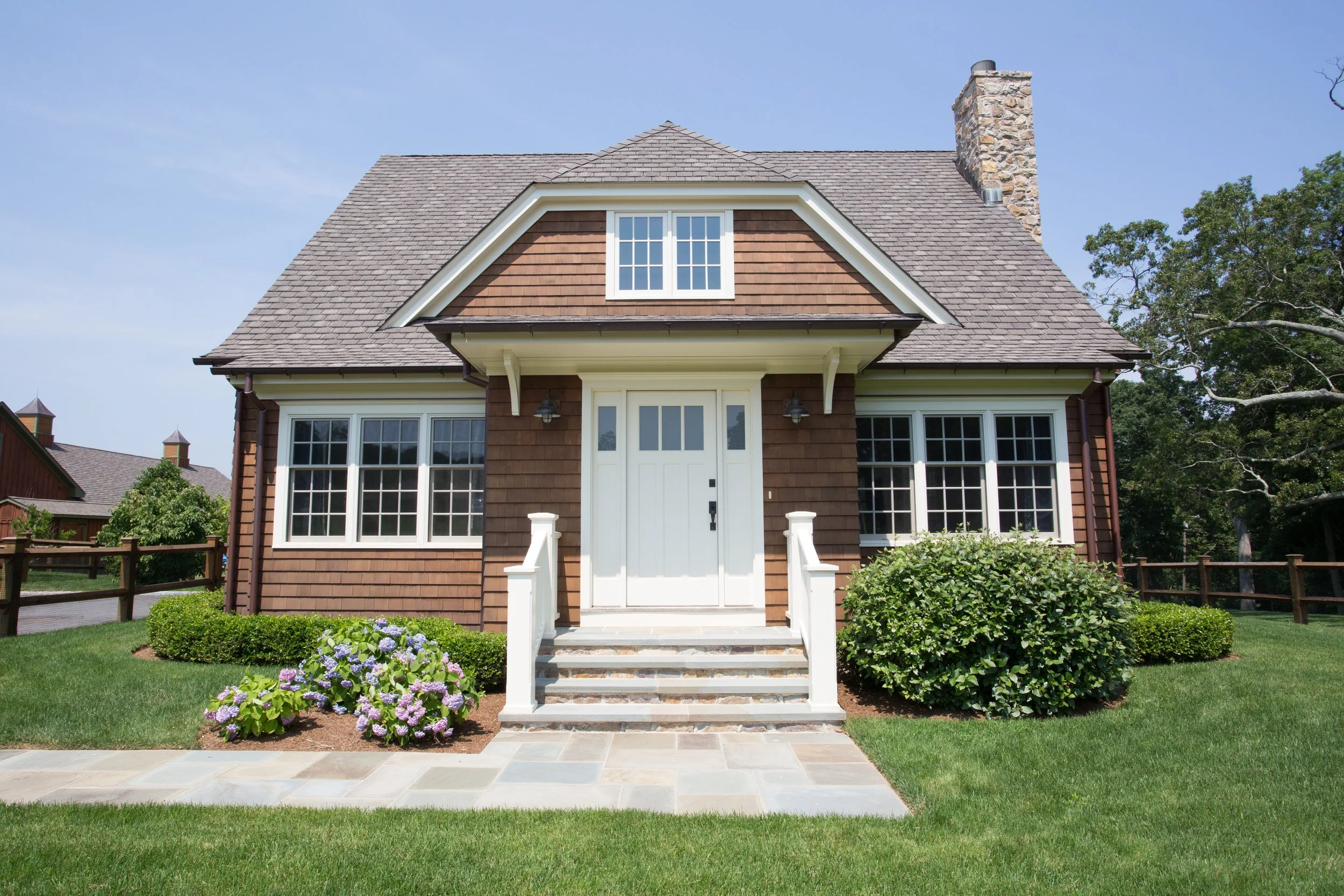 Front view of a house with brown wooden siding, white front door, multiple windows, a stone chimney, and landscaped lawn with bushes and purple flowers.