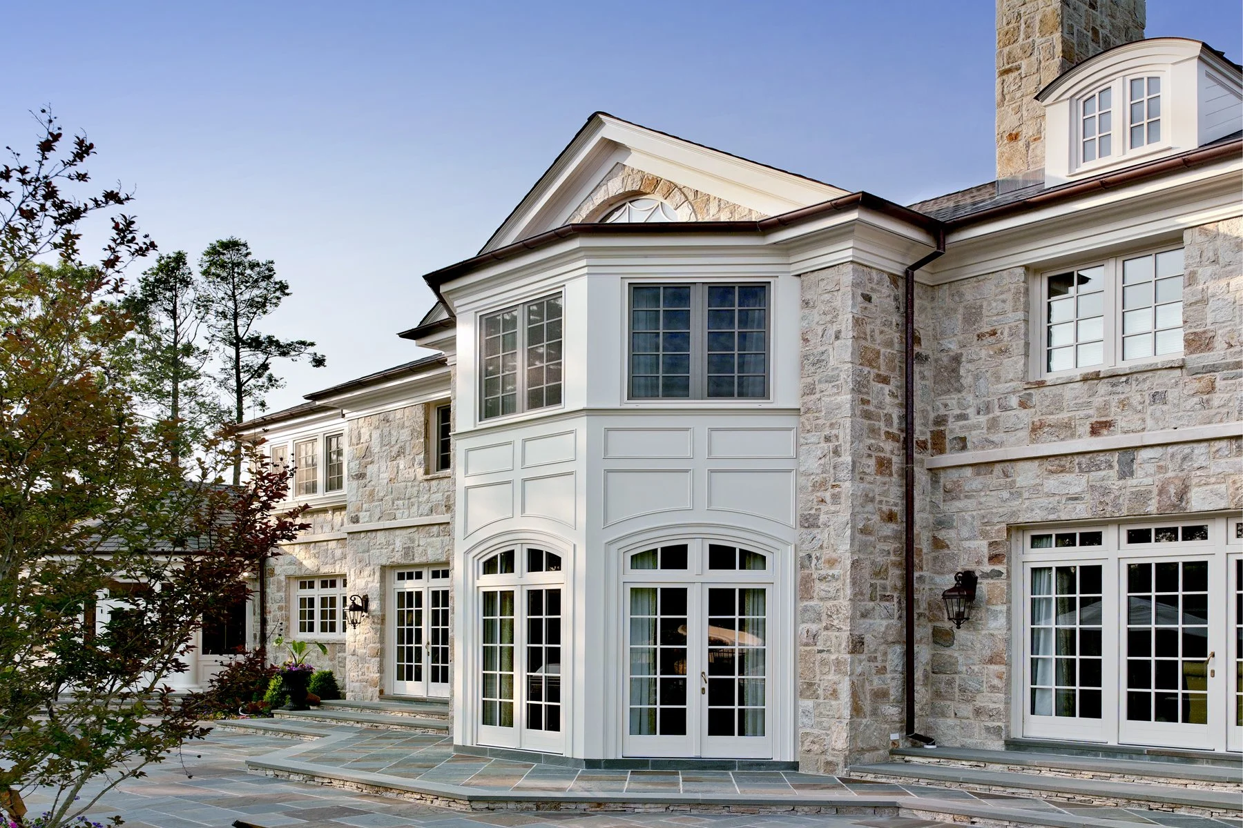 Front view of a large stone and white trim house with multiple windows, including a rounded bay window, and a slate tile patio area.