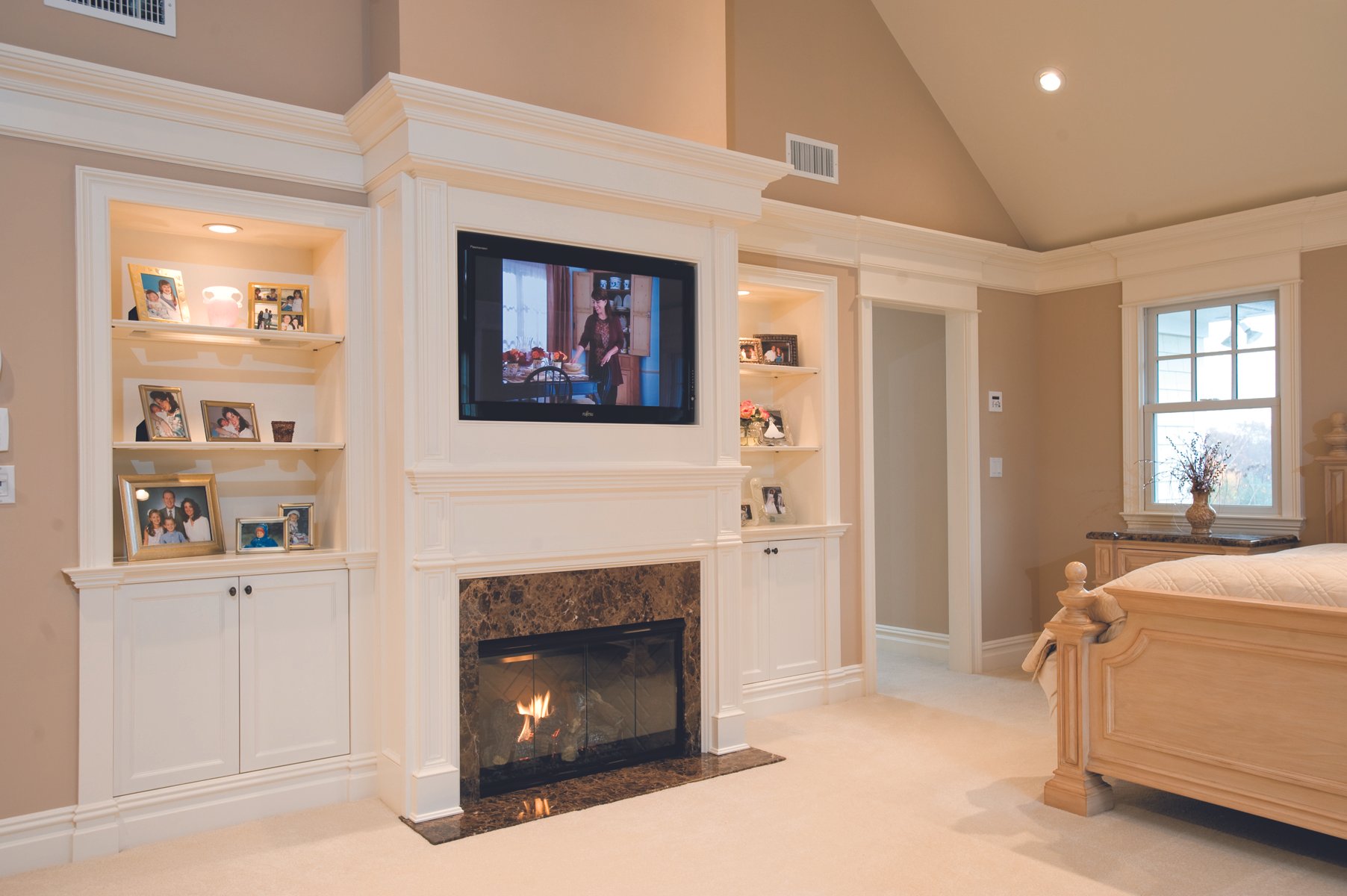 A living room with a fireplace, white built-in shelves with family photos and decorations, a mounted flat-screen TV, a window with a vase on the windowsill, and part of a bed on the right.