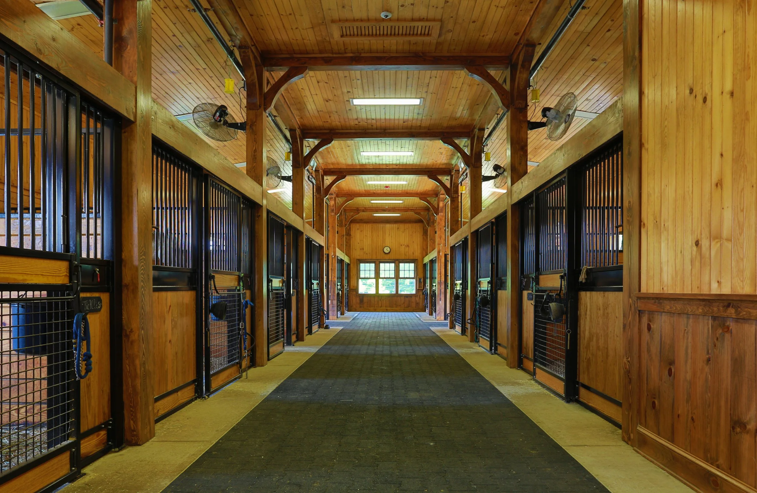 Indoor horse stable aisle with wooden stalls, black gates, and fans attached to the ceiling.