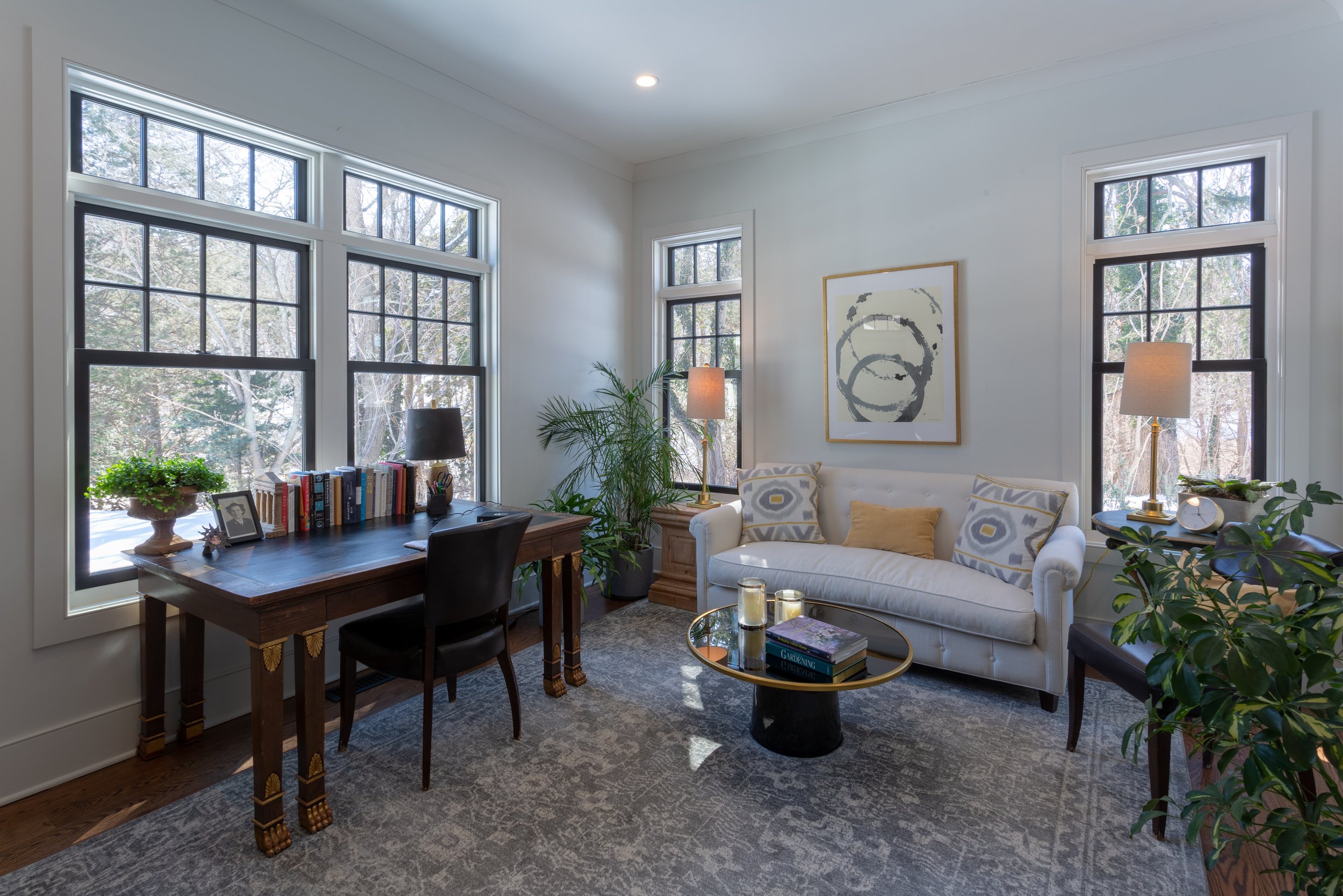 Bright living room with three large windows, a white sofa with patterned and yellow pillows, a round coffee table with candles, a wooden desk with books and a black lamp, and various potted plants.