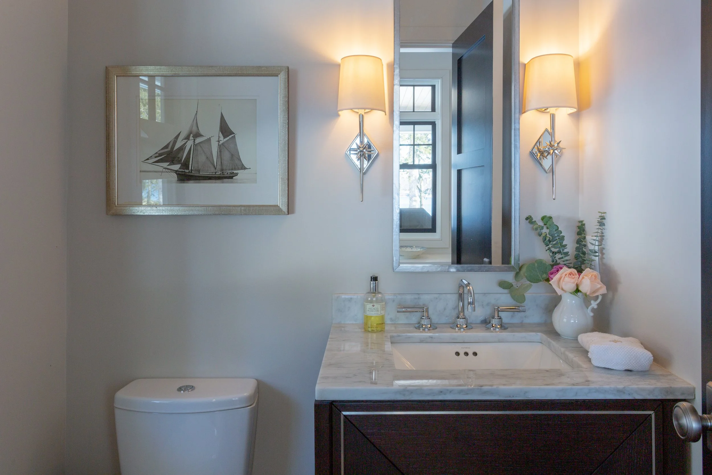 Bathroom with a framed picture of a sailing ship, wall-mounted lamps, a marble countertop sink with a mirror, a vase of pink roses and greenery, a soap dispenser, and neatly folded white towel.