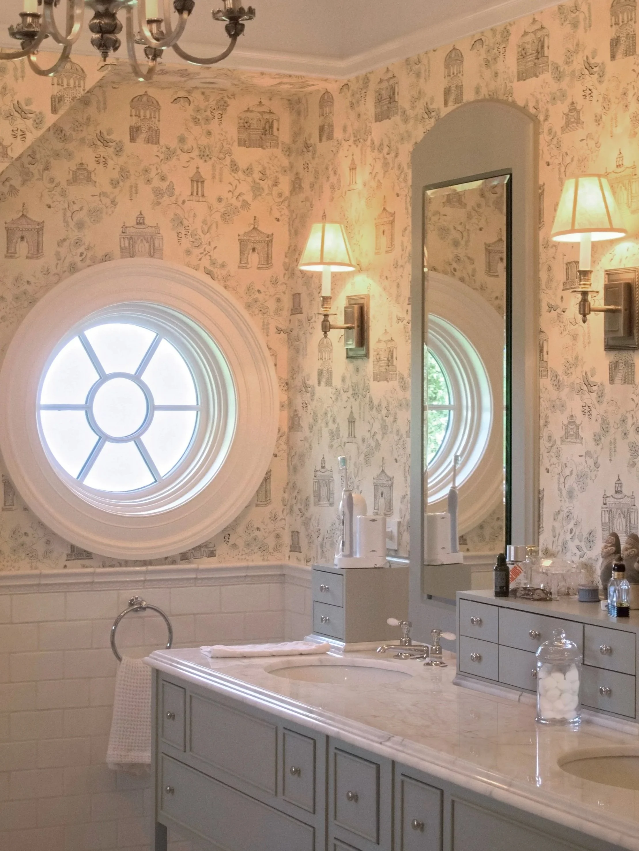 Bathroom with wallpaper featuring architectural drawings, circular window, double-sided mirror, sconces, white vanity with marble countertop, and toiletries.