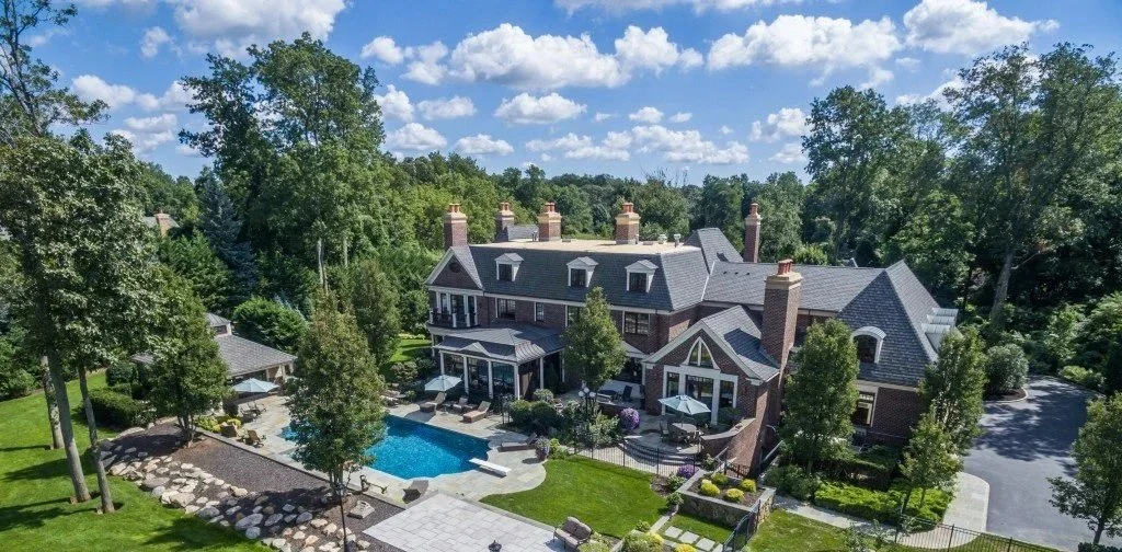 A large, elegant brick house with multiple chimneys, surrounded by lush trees, features a backyard with a swimming pool, patio area with outdoor furniture, and a landscaped garden with a circular patio and pathway.