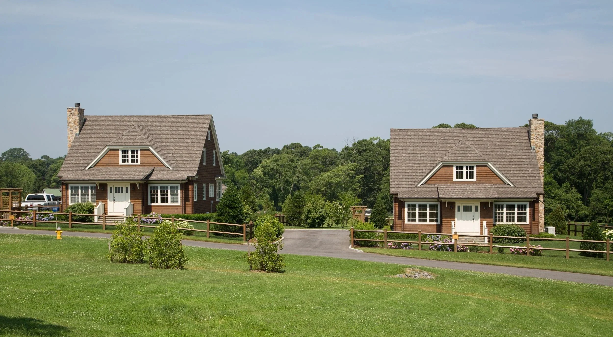 Two brown houses with large front yards and fences, set against green trees under a cloudy sky.