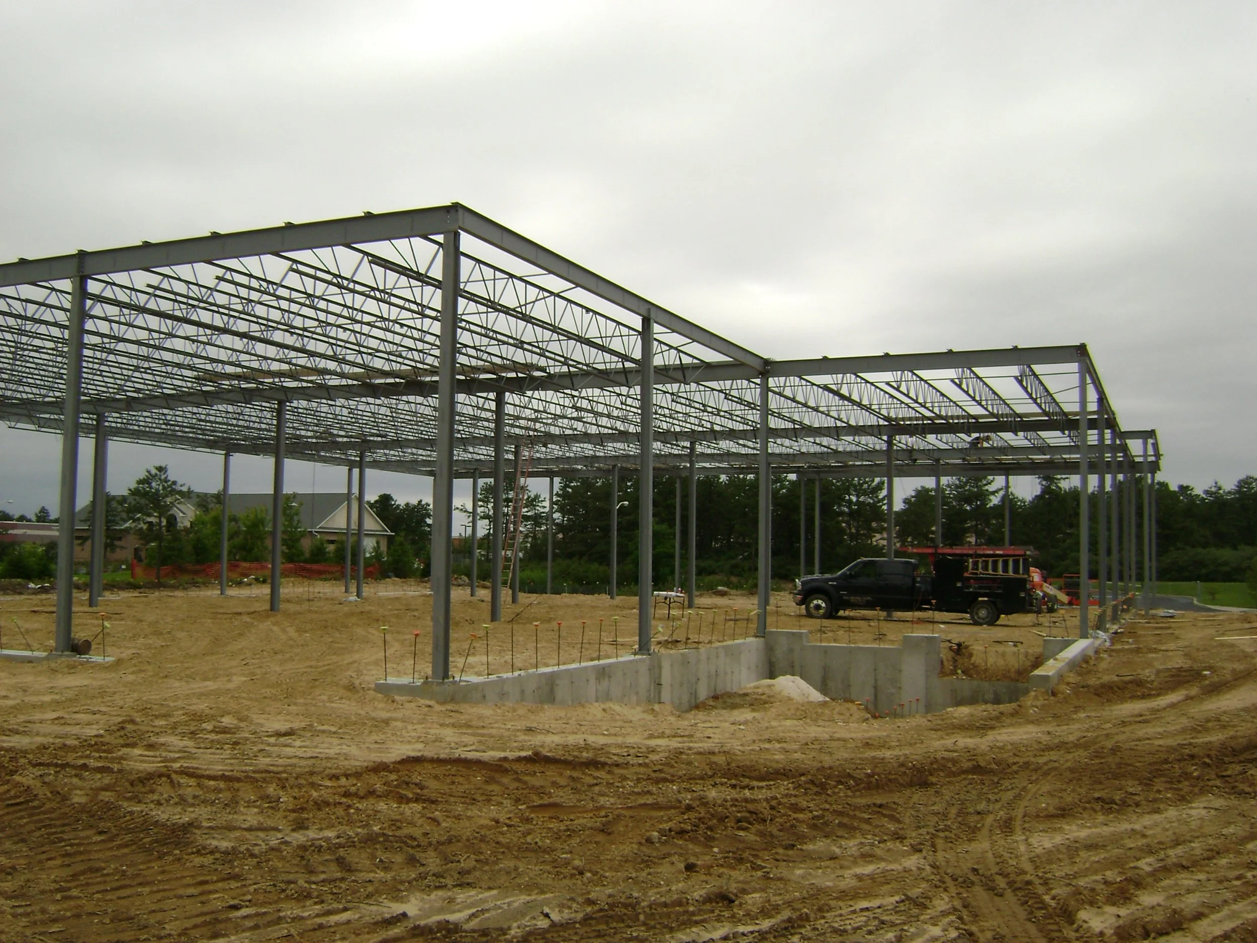 Steel framework for a building under construction on a dirt lot, with cloudy sky and a few houses in the background.