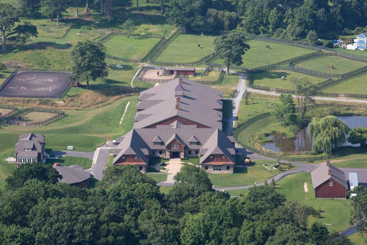 A large horse farm with multiple fenced paddocks, a pond, a main barn with a connecting building, and riding arenas, surrounded by trees and green lawns.