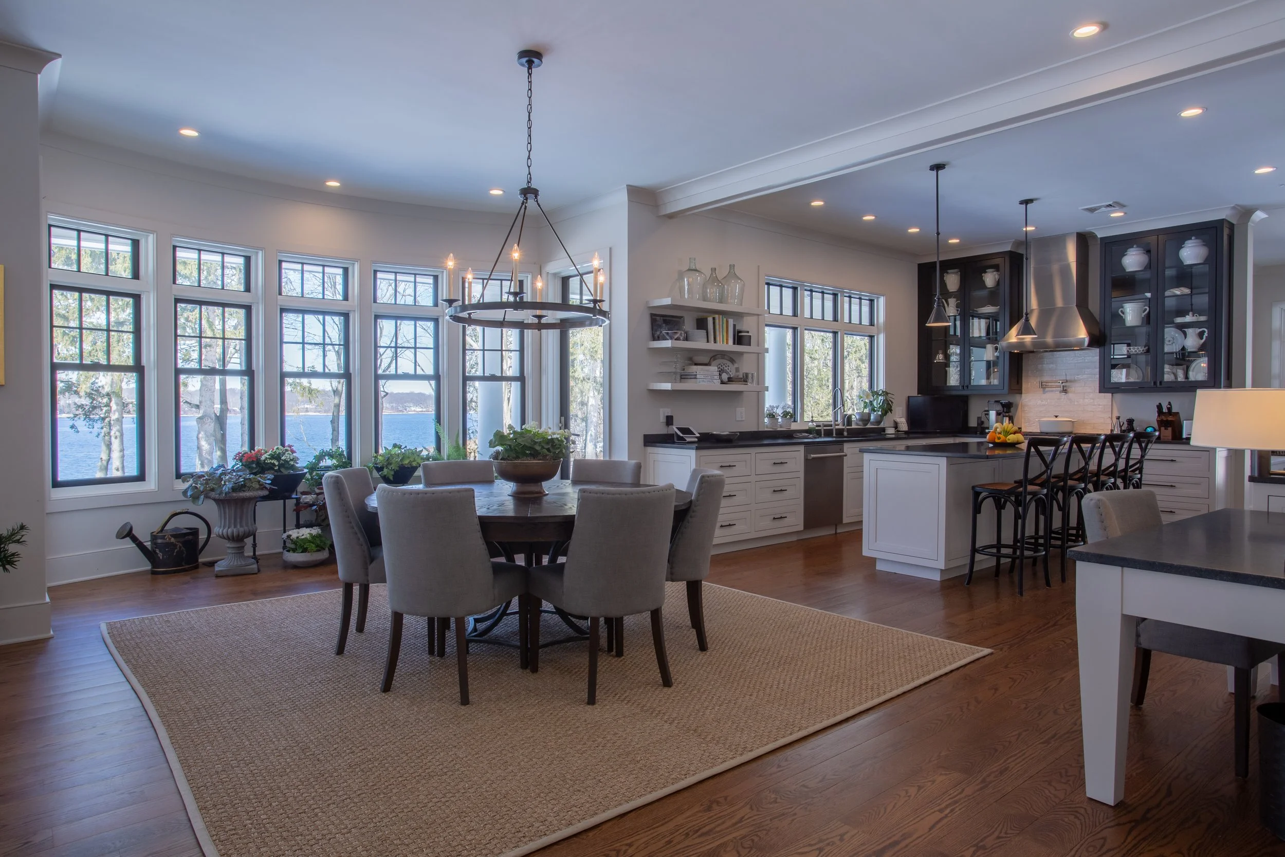 Bright open kitchen and dining area with large windows showing a view of trees and water. Contains a round dining table with eight gray upholstered chairs, a chandelier above, and a kitchen with white cabinets, black countertops, and a stainless stee
