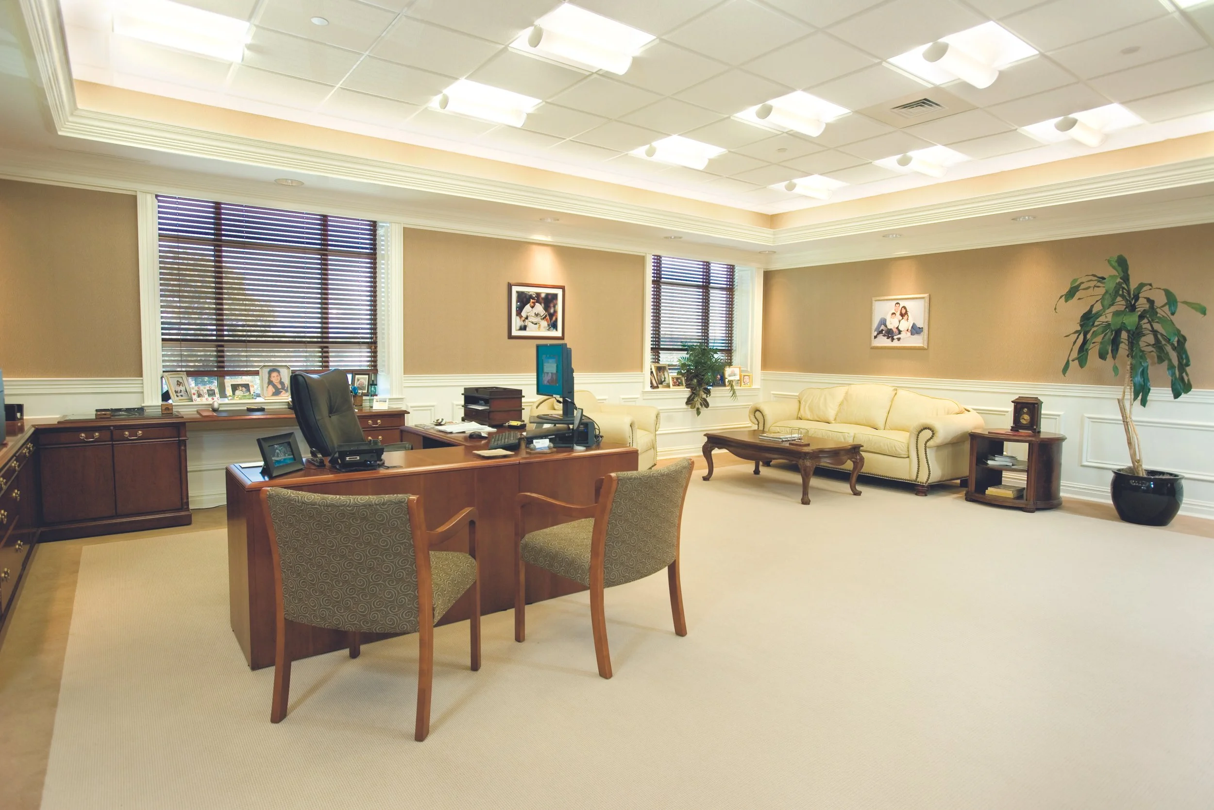 Spacious office with large desk, multiple chairs, cream-colored sofa, wooden coffee table, potted plant, framed photos, and artwork, illuminated by ceiling lights and window blinds.