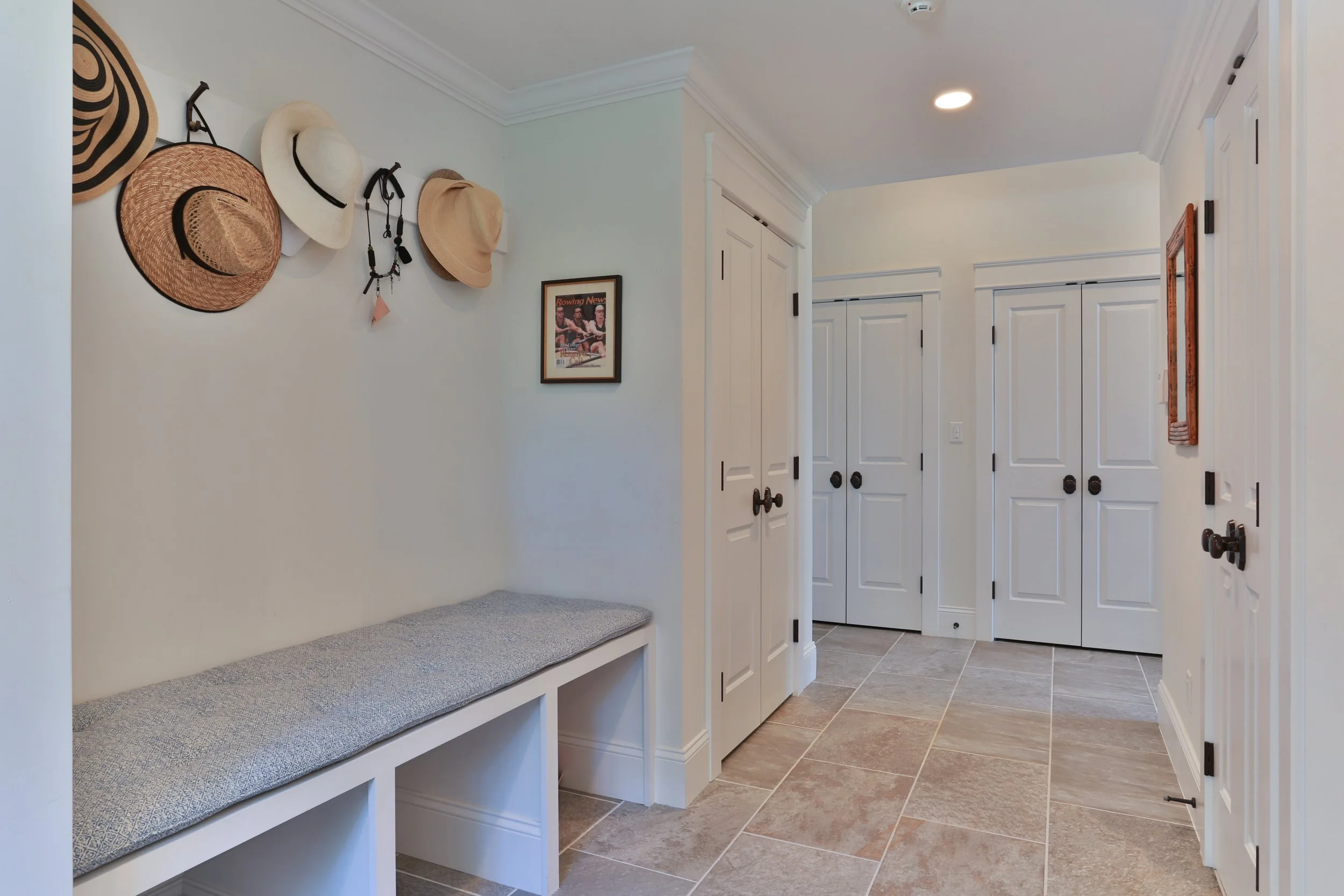 Entryway with white shelving and hanging hats, framed picture on wall, tiled floor, and white doors.