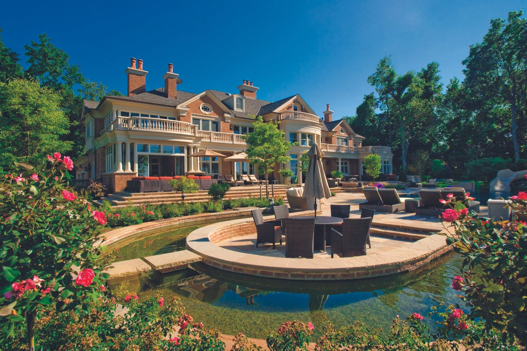Large, luxurious brick house with multiple chimneys, spacious balcony, and large windows, surrounded by lush green trees. In the foreground, there is a landscaped backyard with a patio that includes outdoor furniture, a small pond with pink flowers, 