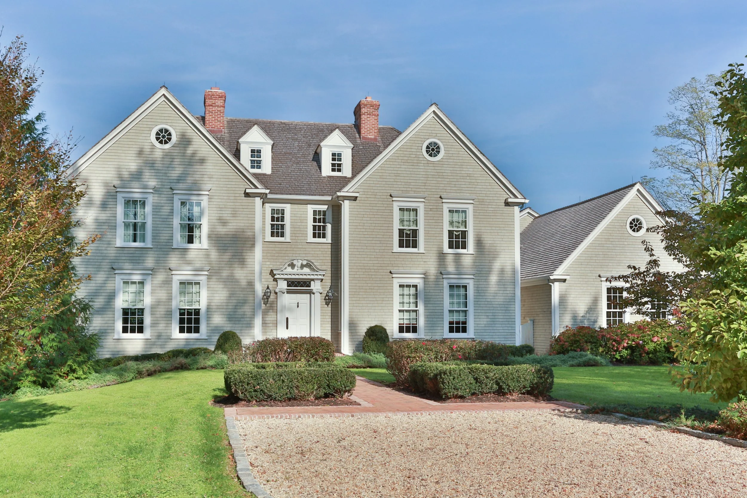 Front view of a large, two-story house with beige siding, multiple windows, a brick pathway, and a well-maintained lawn with shrubbery and trees.