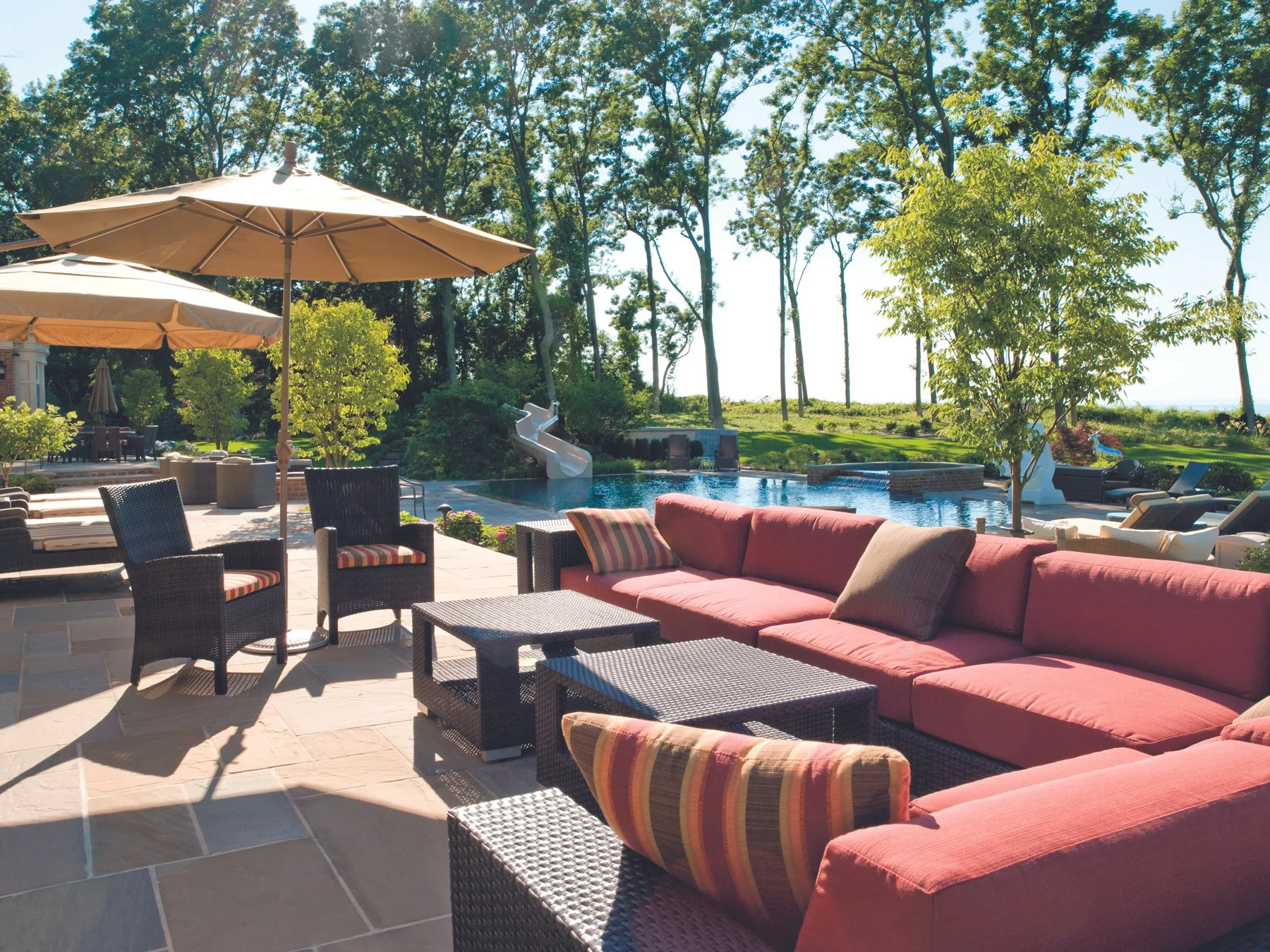 Spacious backyard with outdoor seating, umbrellas, swimming pool, slide, and lush greenery with trees