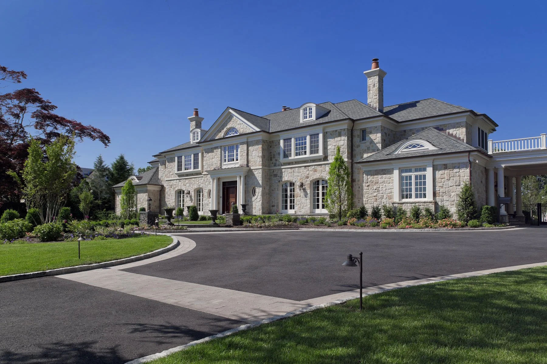 Large, elegant stone mansion with multiple chimneys, surrounded by landscaped gardens and a paved driveway under a clear blue sky.