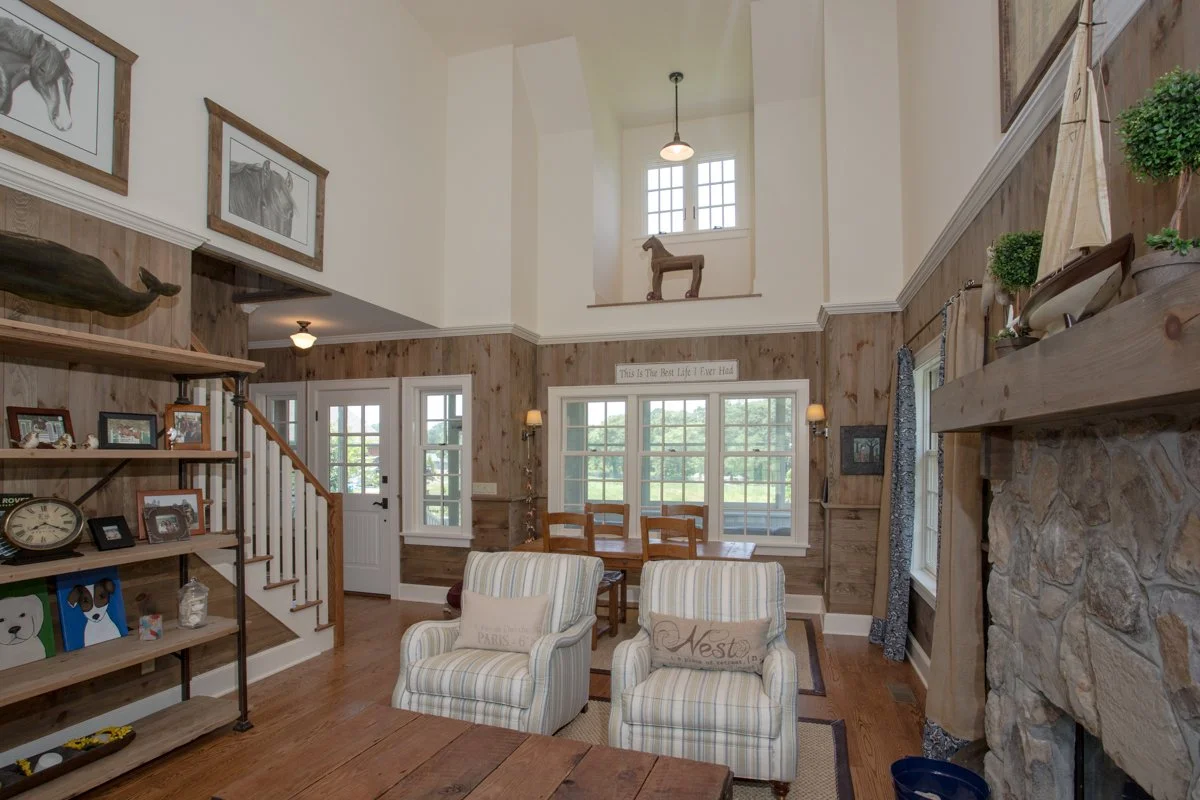 Living room with wooden wall paneling, two striped armchairs, a dining table with chairs, a fireplace with stone surround, and decorative items like framed photos, a whale sculpture, and ornamental plants.