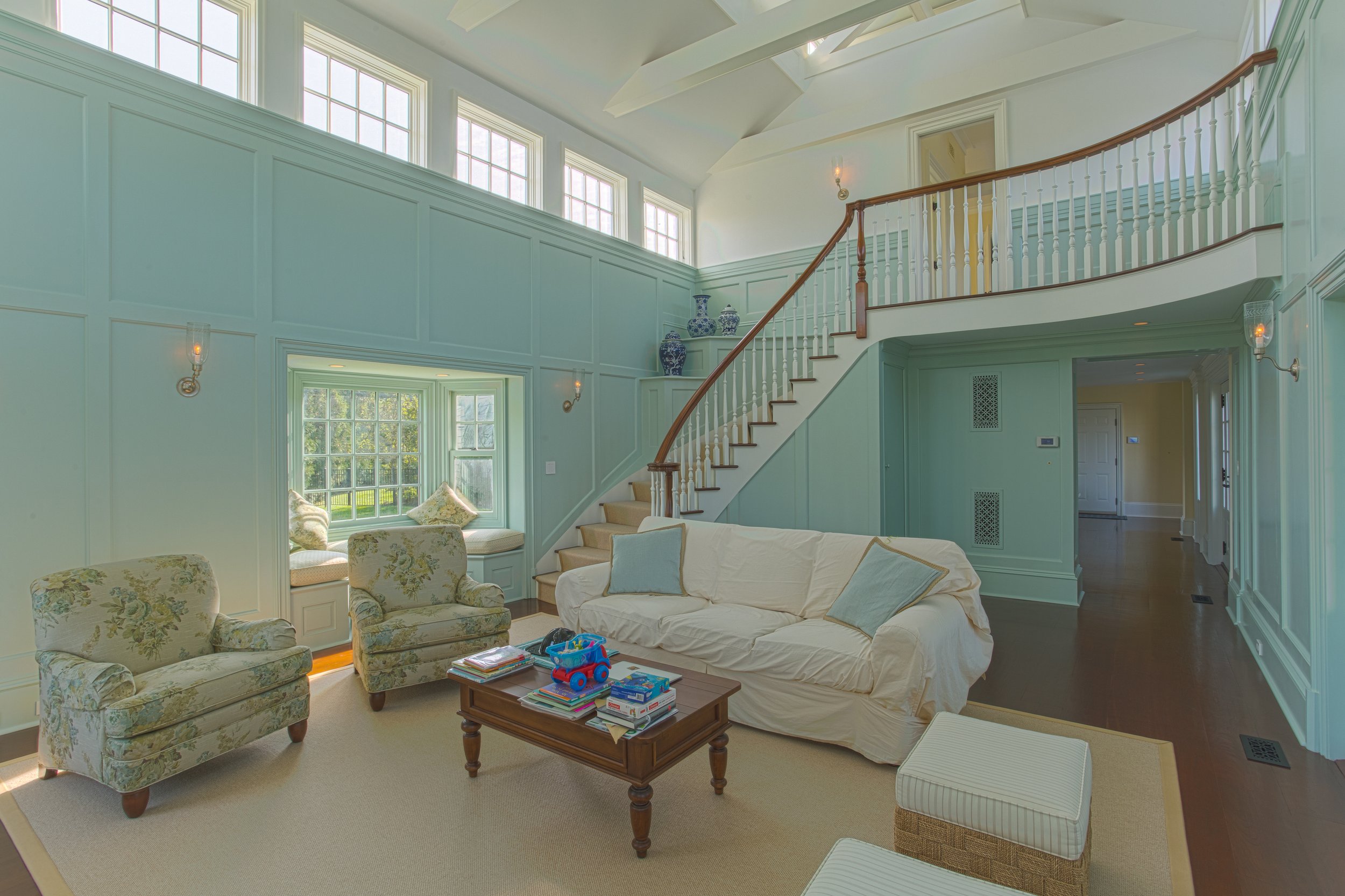 Living room with high ceilings, large windows, and a curved staircase. Contains floral armchairs, a white sofa with pillows, a coffee table with books and toys, a window seat with cushions, and decorative blue and white vases.