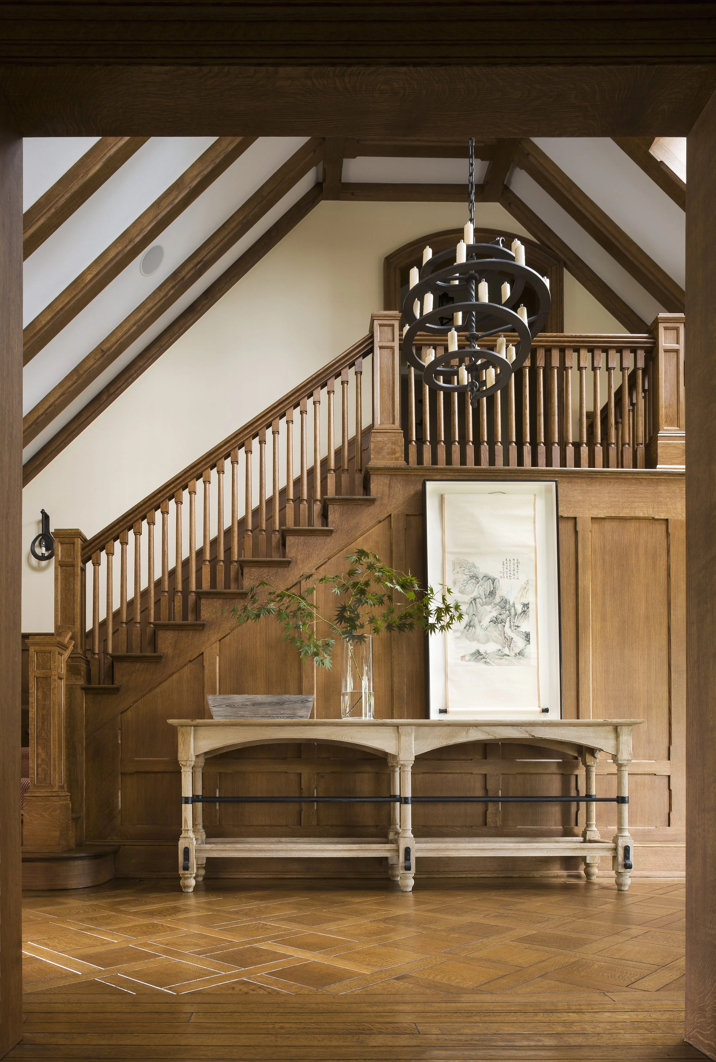 Interior view of a wooden staircase with a balcony, a hanging chandelier, a console table with a potted plant, a framed artwork, and wood-paneled walls and ceiling.